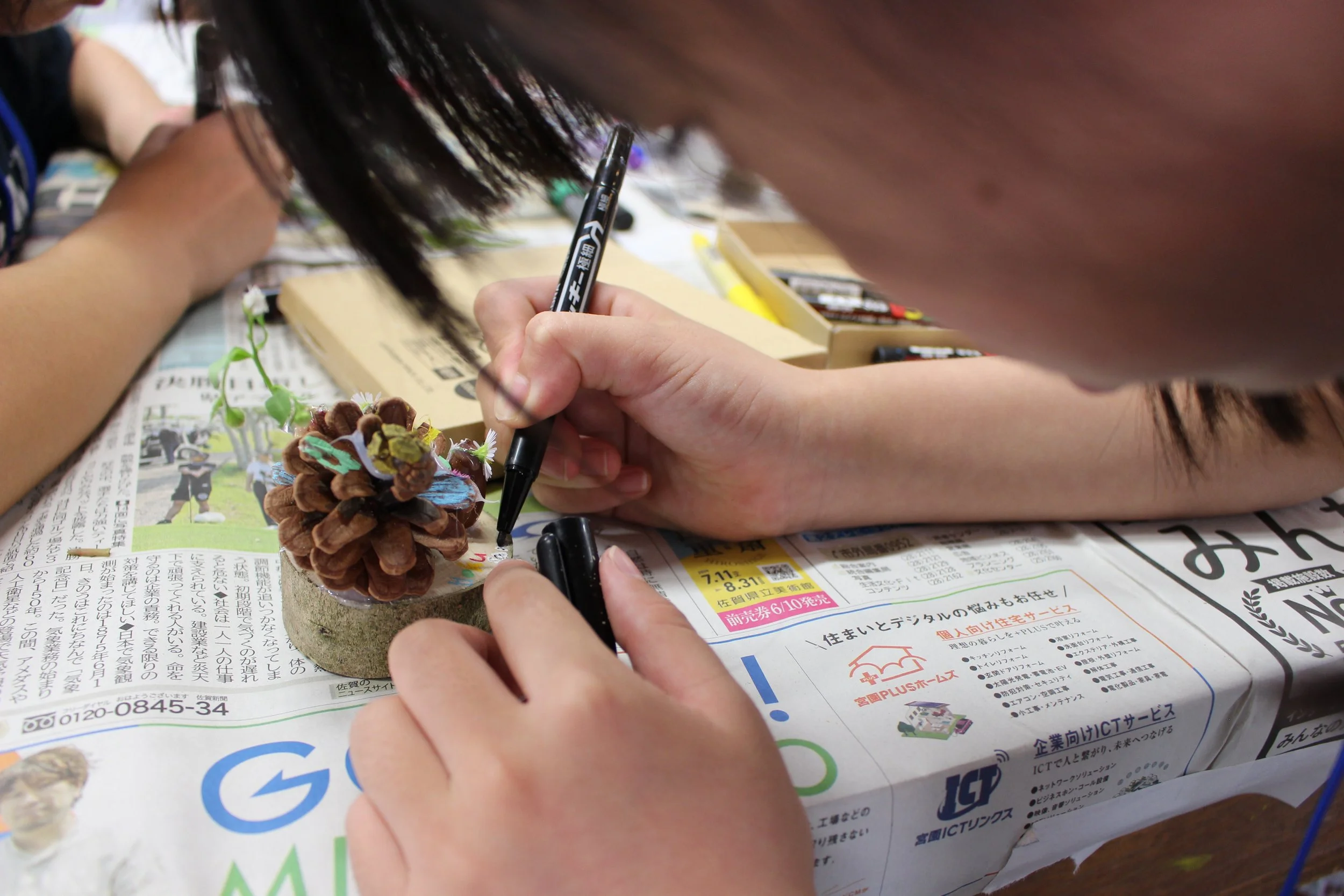 人がペンを使って木の実の模型に絵を描いている様子。テーブルには新聞紙と画材が置かれている。