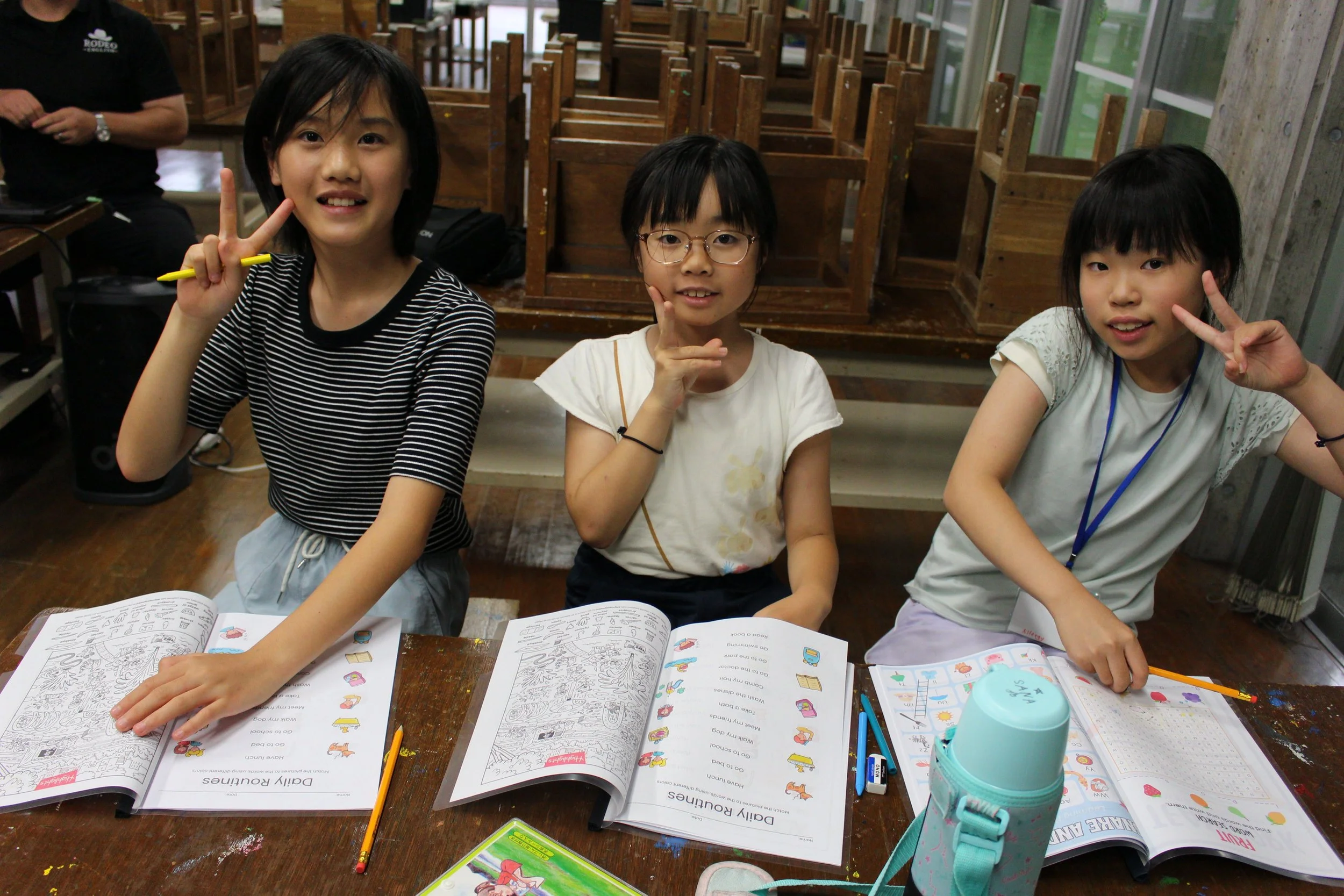 三人の女の子が机に座っている教室の様子。彼女たちは色鉛筆や本を持ち、ポーズをとっている。