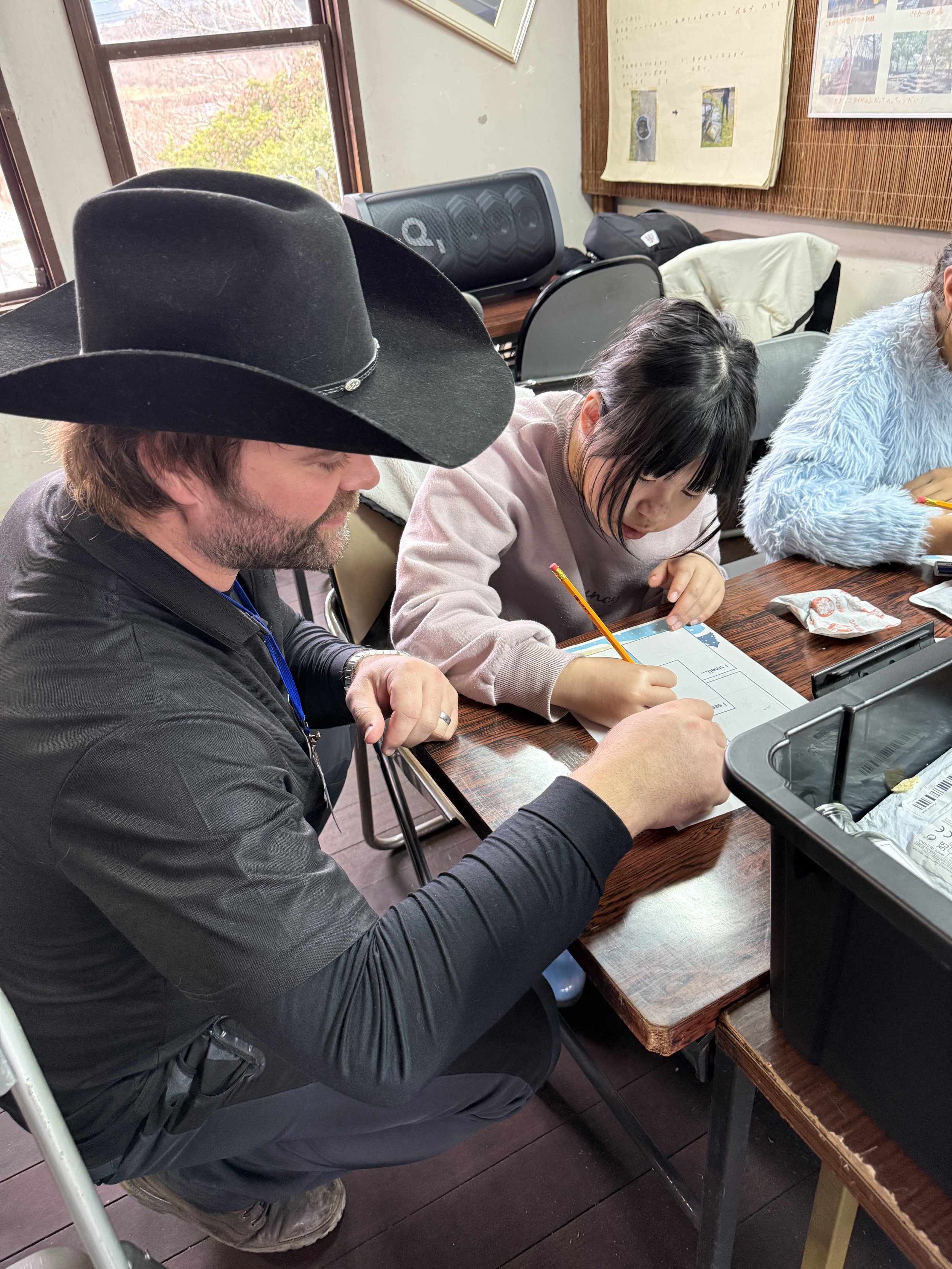 大人と子供がテーブルで座りながら一緒に書き物をしている。大人は黒い帽子をかぶっている。室内で自然光が入る。