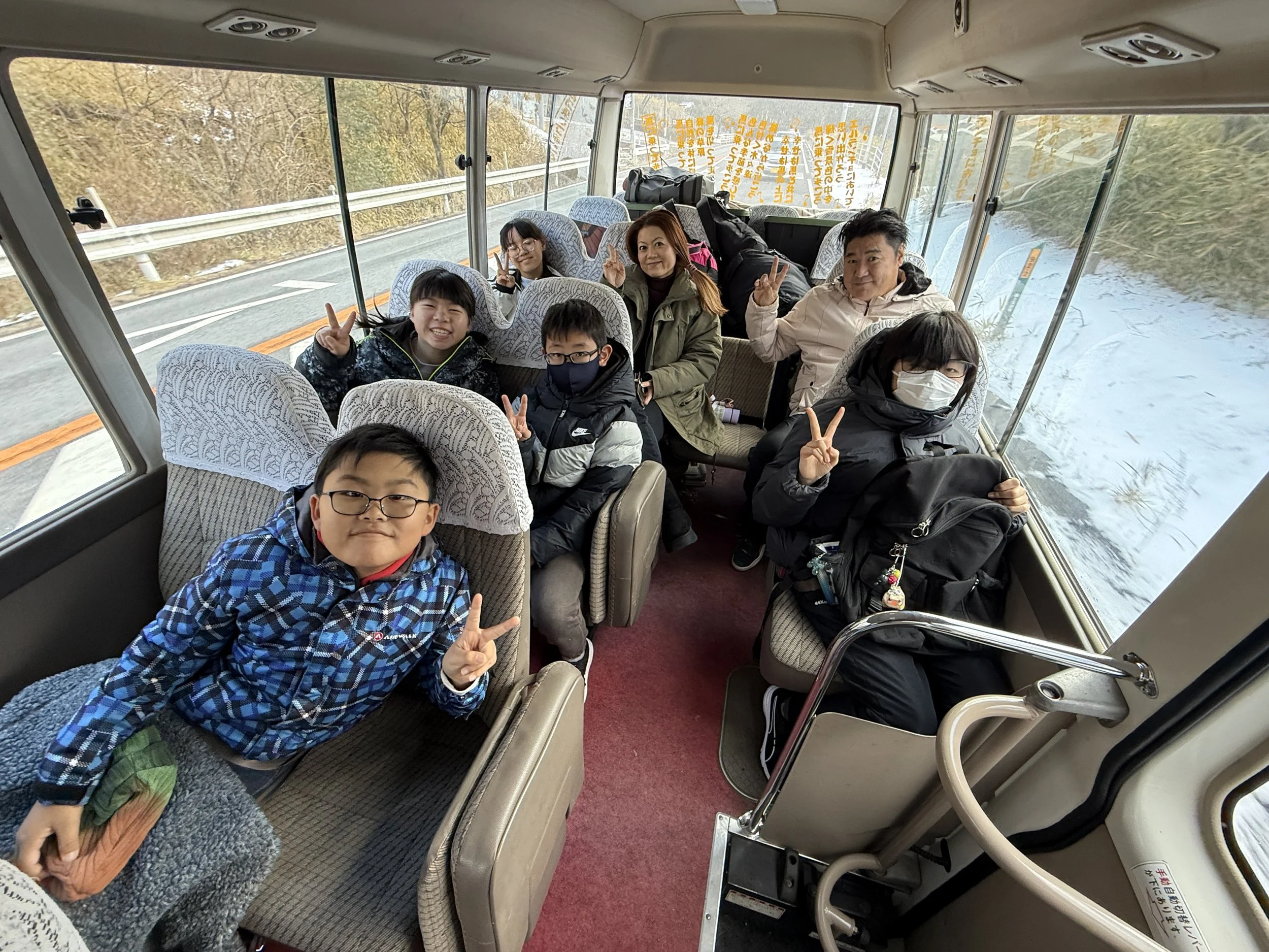 バスの中で座る大人と子供たちが笑顔とピースサインをしている。車内は窓の外に冬の景色が見える。