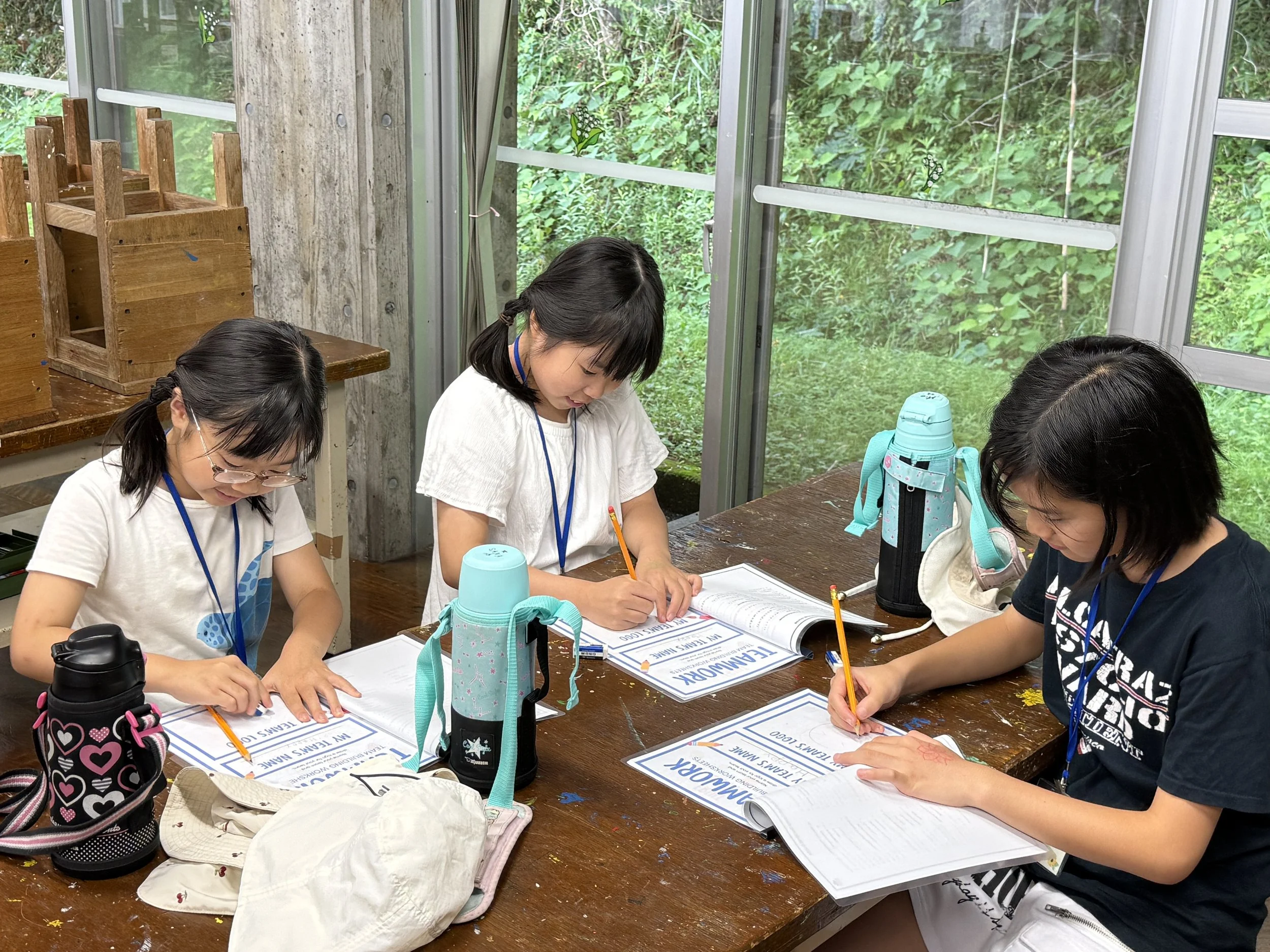 室内の木製のテーブルに座る3人の少女が、教科書とノートに記入している。背景には大きな窓越しに緑豊かな自然が見える。テーブルには水筒やバッグも見える。