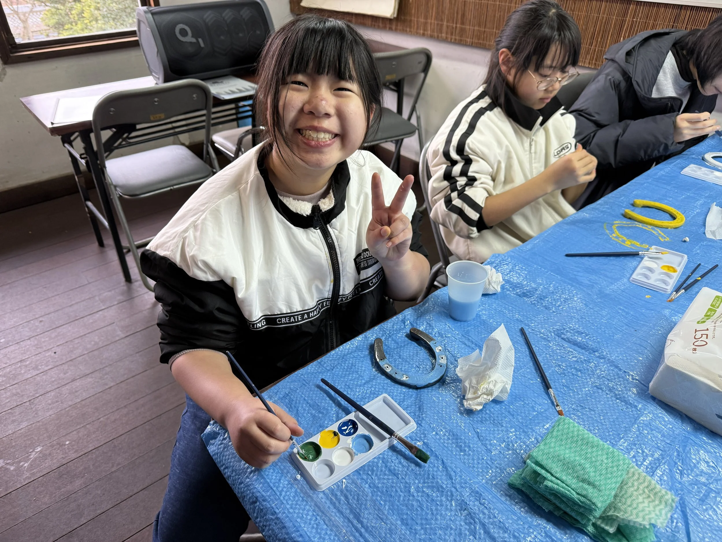 子供たちが絵の具でペイントをしているテーブルの横に座っている女の子が笑顔でピースサインをしている姿。