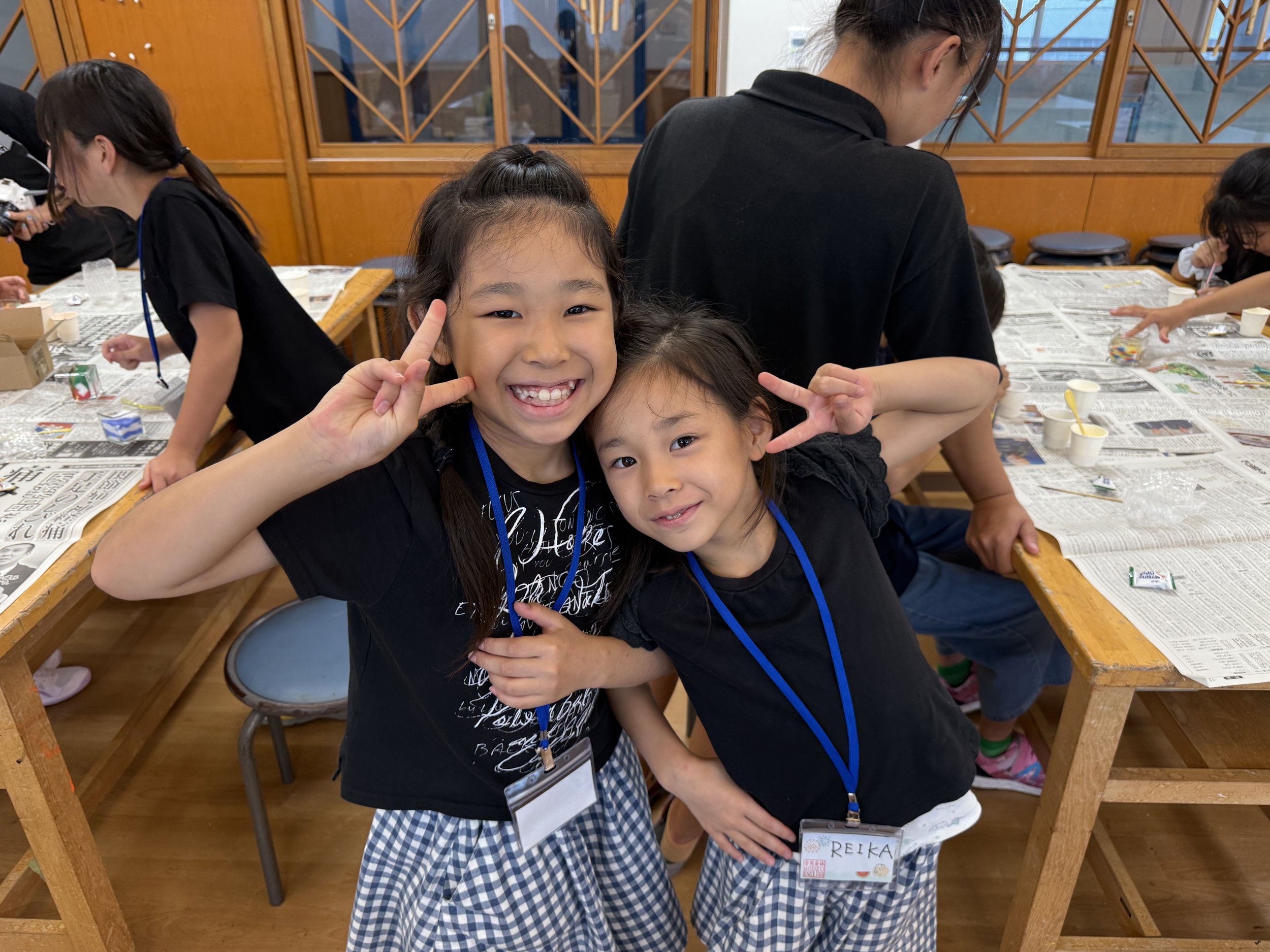 笑顔の二人の女の子がポーズをとっている。背景には、新聞紙を敷いた長い木のテーブルと他の子供たちがいる室内の様子が見える。