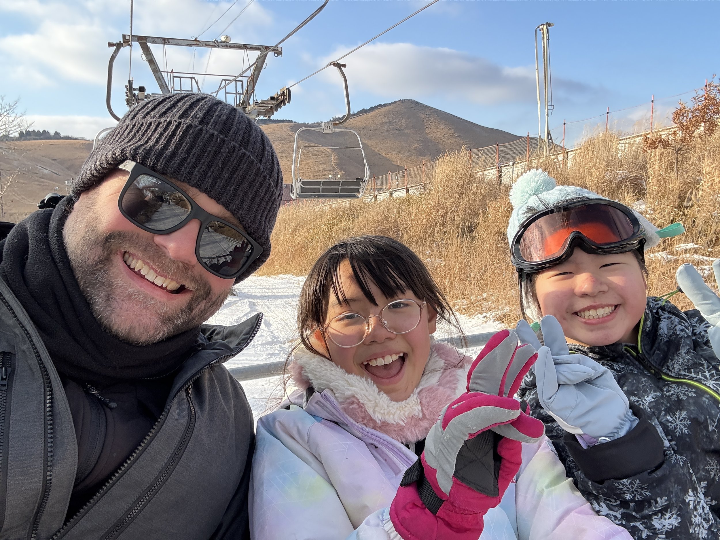 雪山の背景にスキーを楽しむ家族の写真。男女二人の子供と男性が笑顔でカメラを見ている。三人とも冬のスキーウェアを着用し、手を振っている。