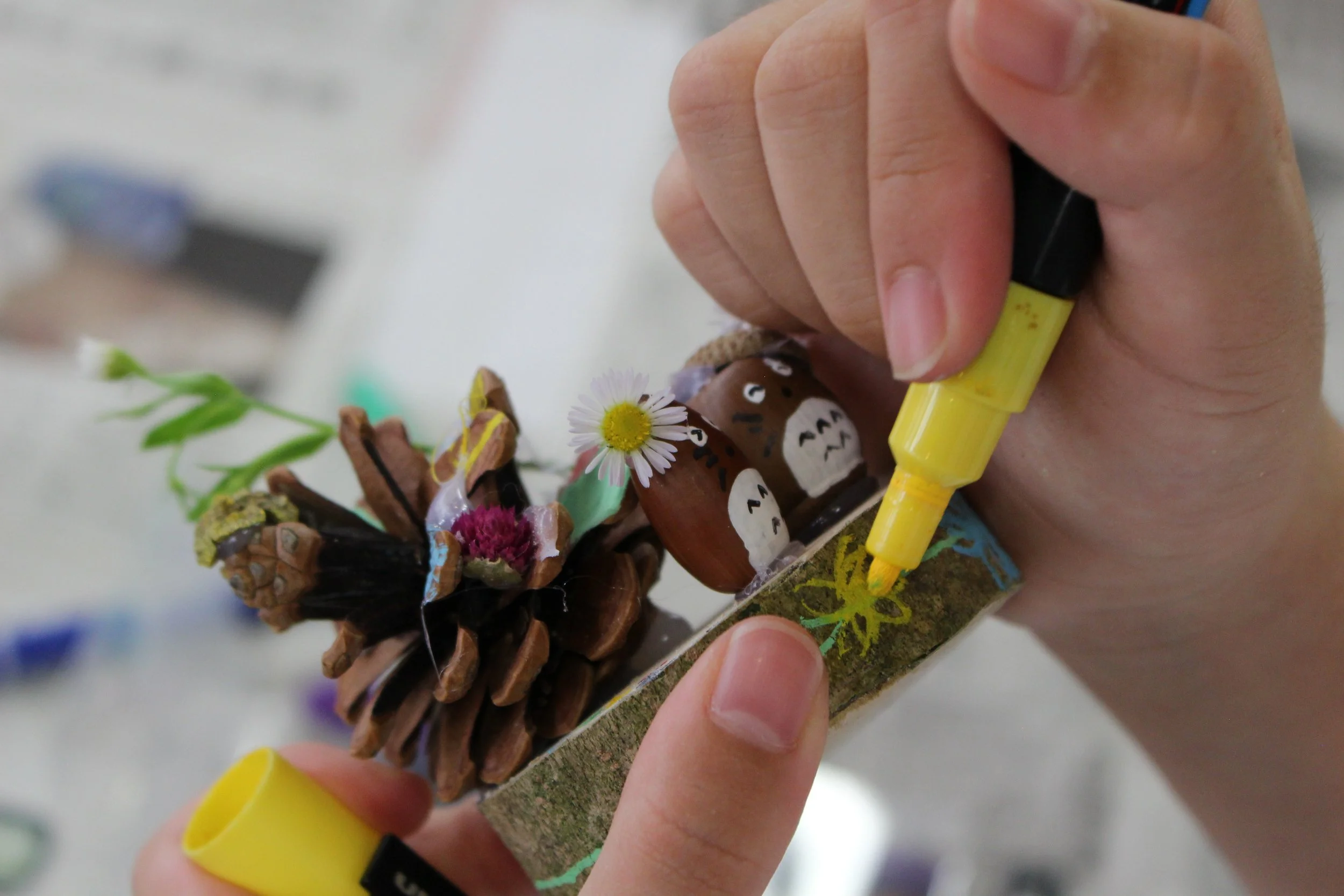 小さな木の実に描かれたキャラクターや花の飾りに黄色のペンで絵を描いている手