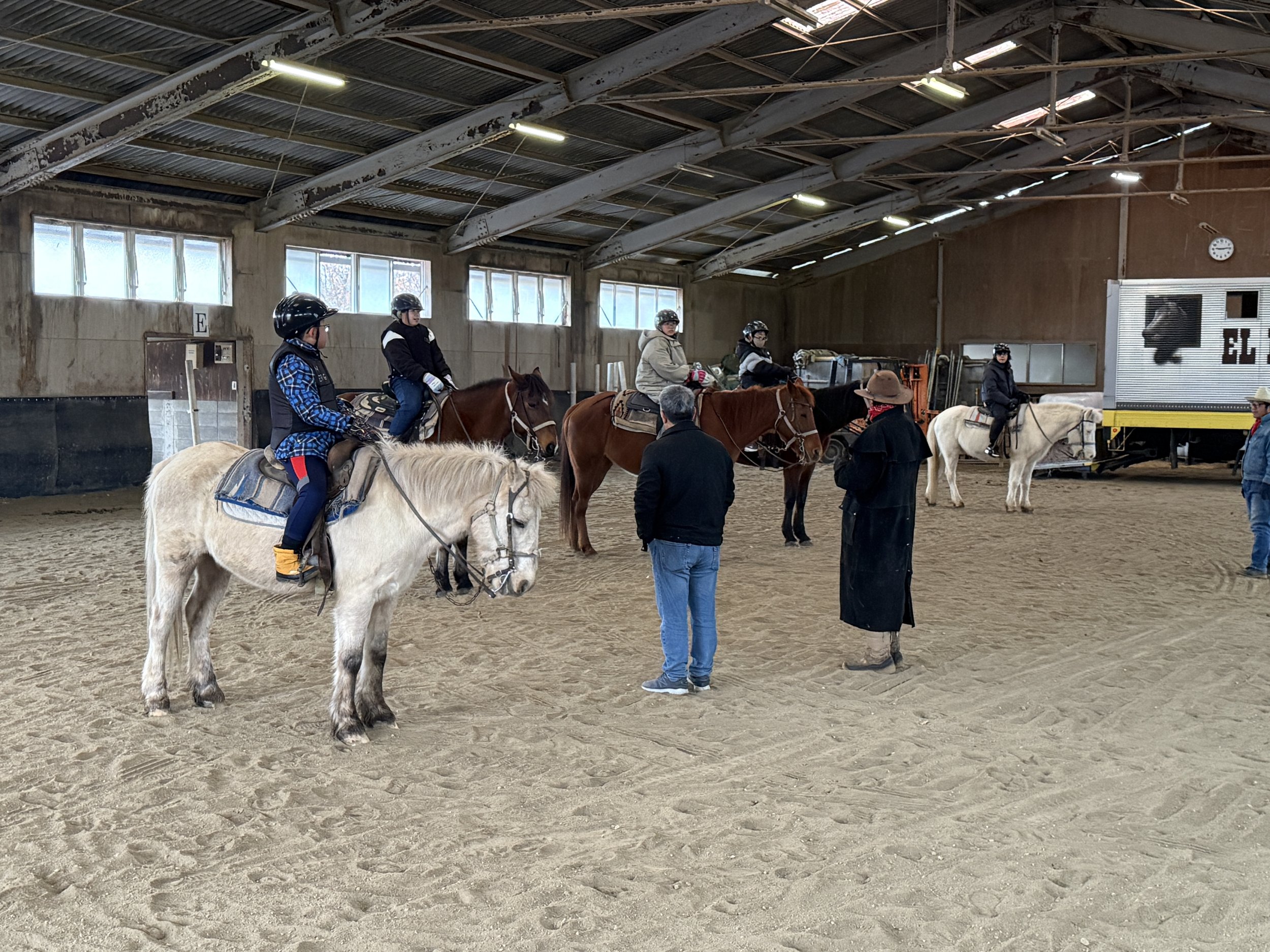 馬場で馬に乗る人たちがレッスンを受けている。インストラクターと話す人もいる。屋内の馬術練習場の様子。