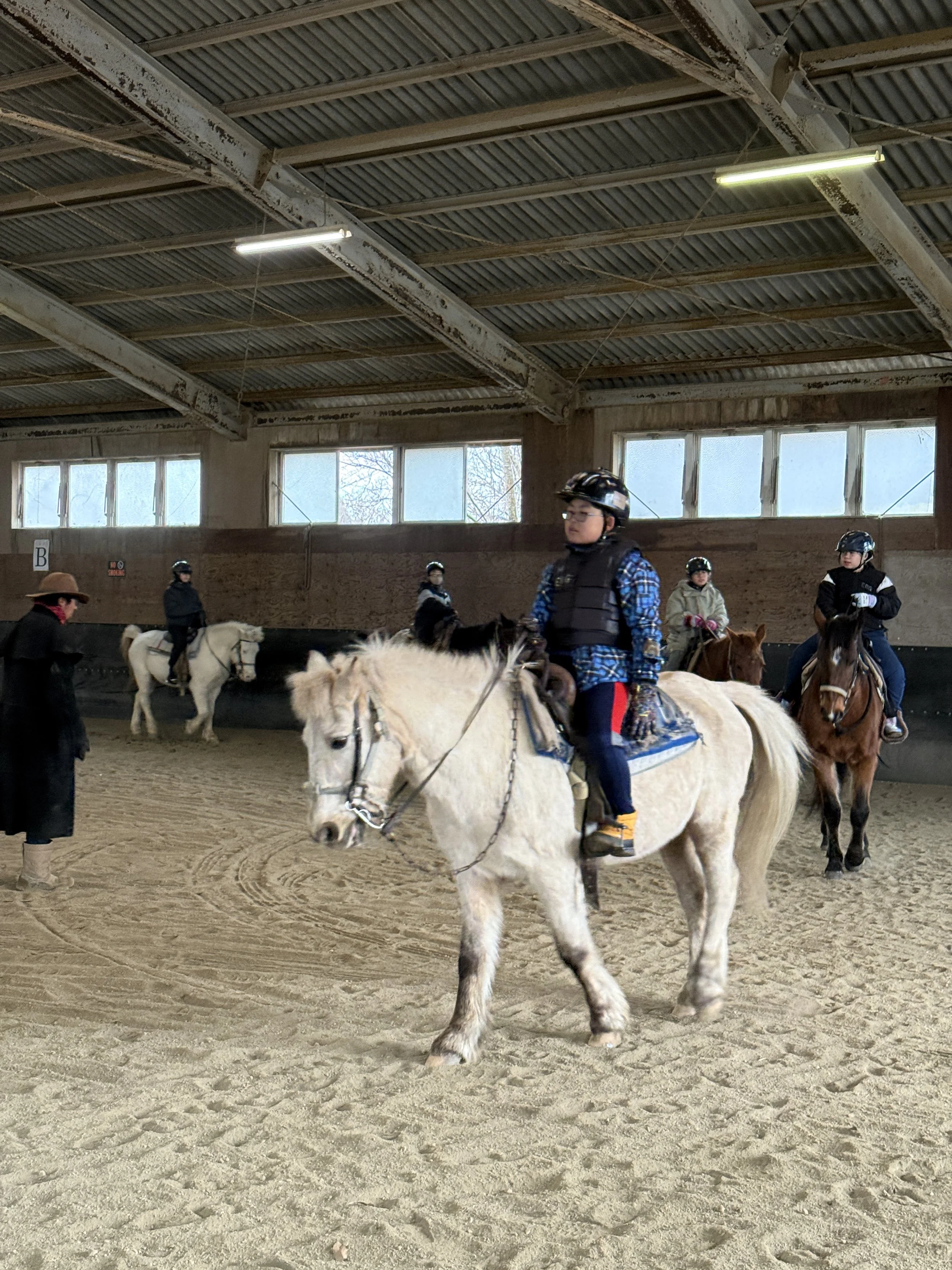 屋内の馬場で馬に乗る人と見学者のグループ。馬に乗っている人はヘルメットを着用している。