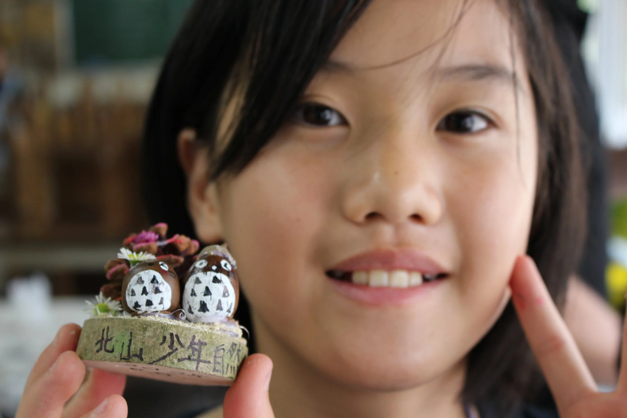 子供が指を指しながら笑顔で木でできた飾りの前で写真を撮る。飾りはカボチャの形をしていて、上にはピンクや白の花と黒い目や鼻、口の顔が描かれた飾りがついている。