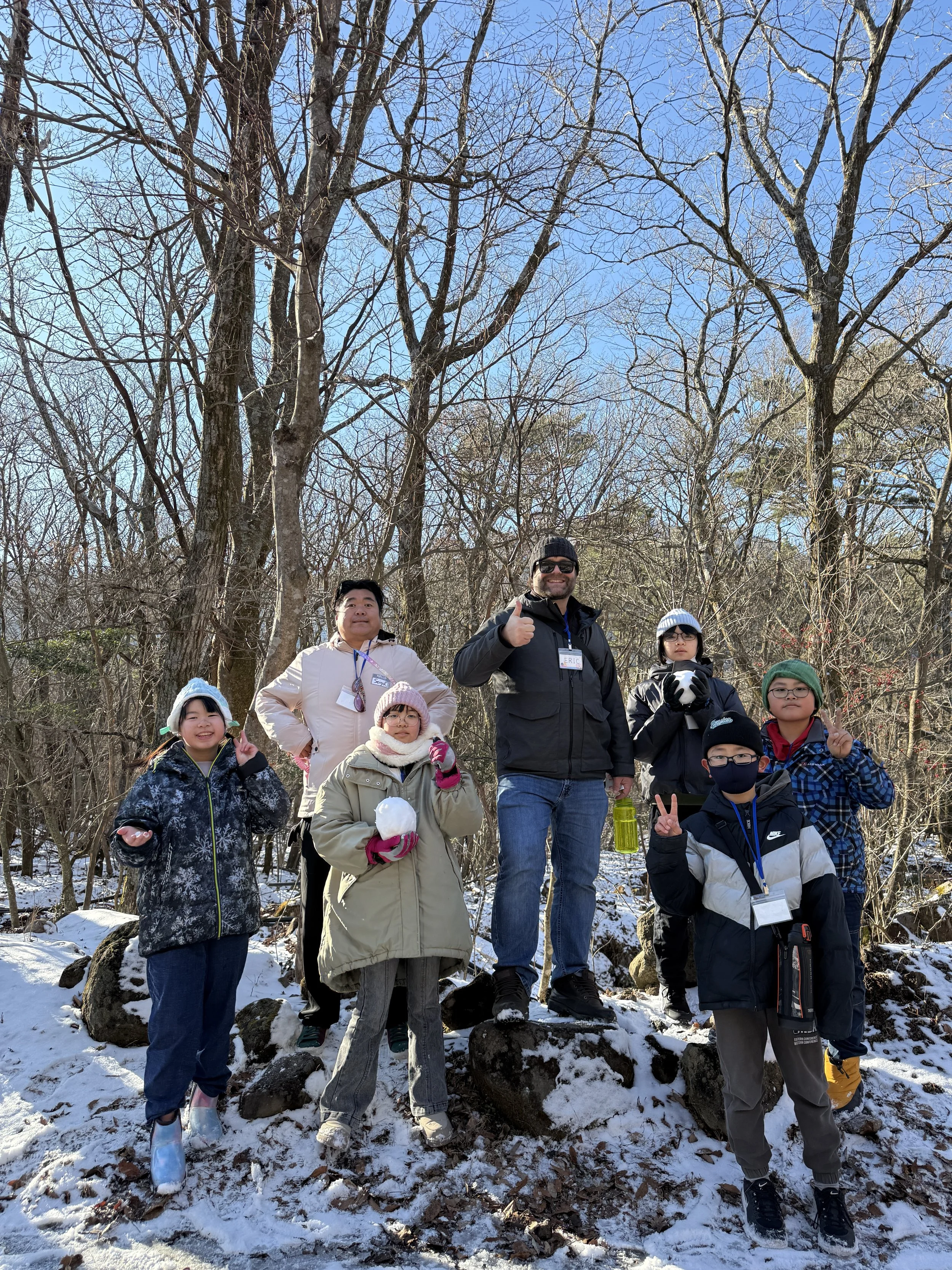 冬の森林の中で、子供と大人のグループが雪の上に立っている。子供たちは防寒具を着て、雪だるまやピースサインをしている。大人もピースやサムズアップのジェスチャーをしている。