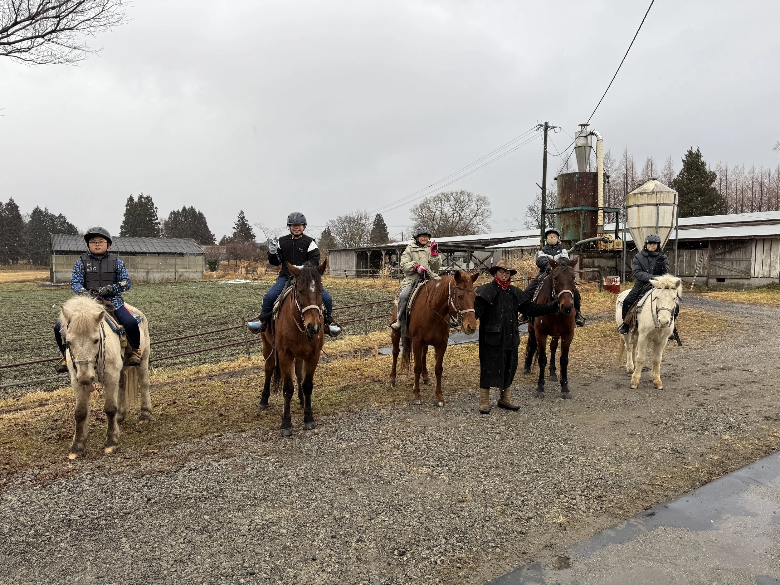 馬に乗った6人の人々と馬乗りの2人のスタッフがいる牧場の風景。背景には農場の建物や木々が見える。