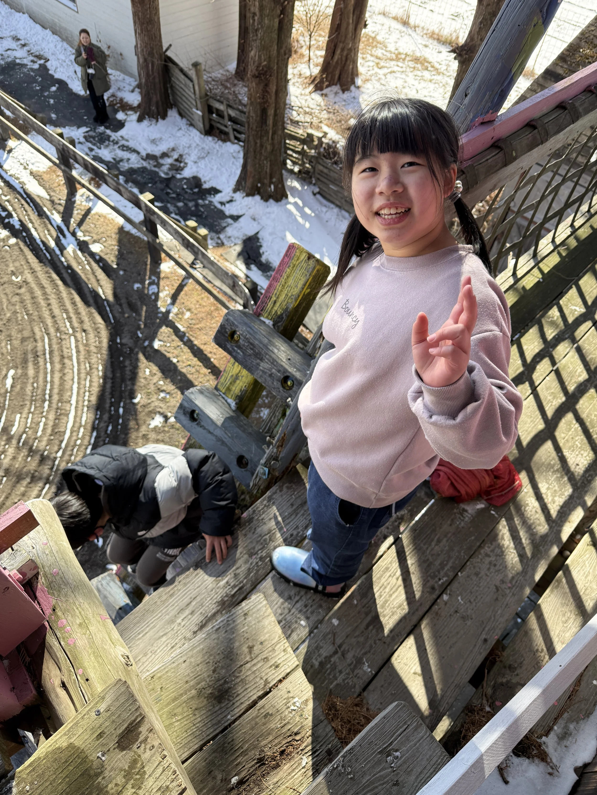 笑顔の女の子が木製の階段の上に立っている。周囲は雪と木々に囲まれ、背景には大きな木と、階段を下る子供。