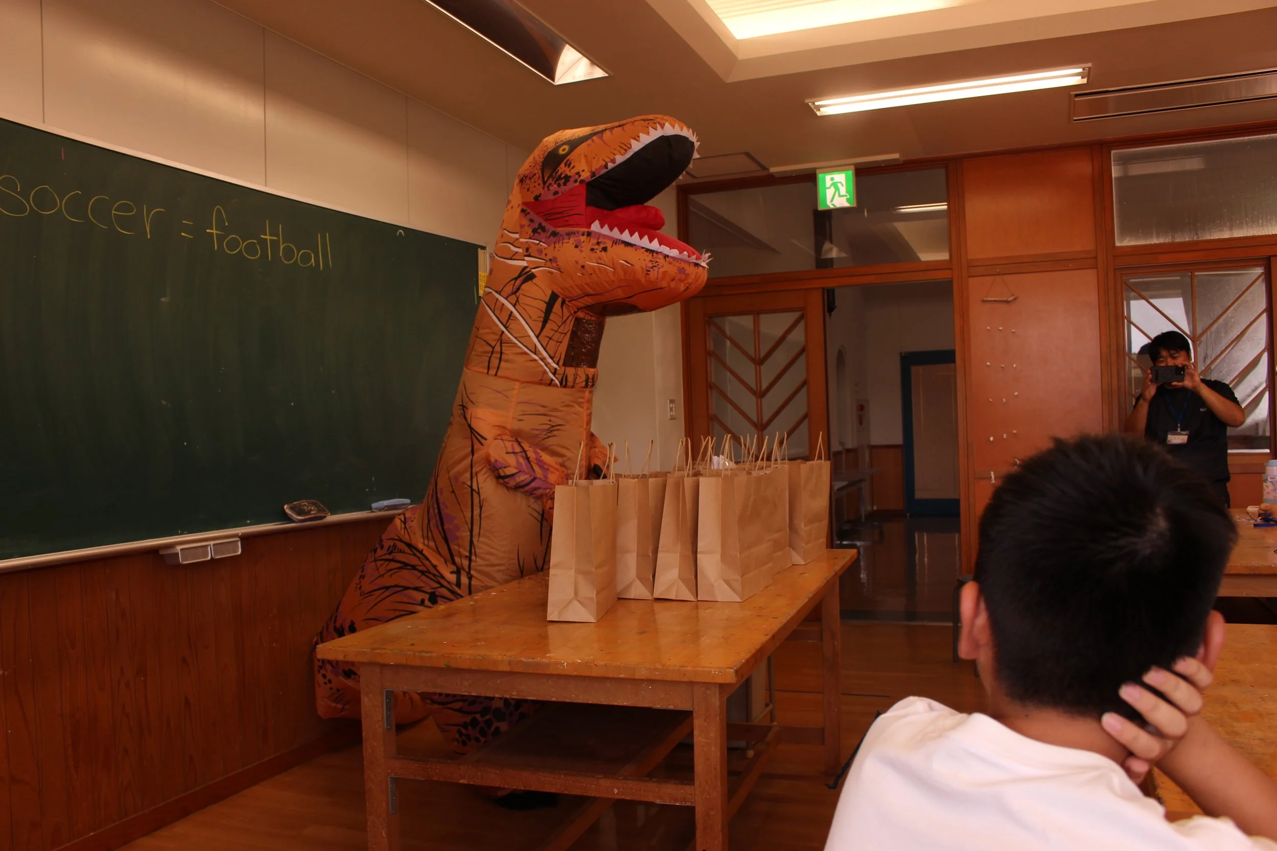 恐竜の着ぐるみを着た人が教室で紙袋の山の前に立っている。教室には黒板と子供たちが座っている。