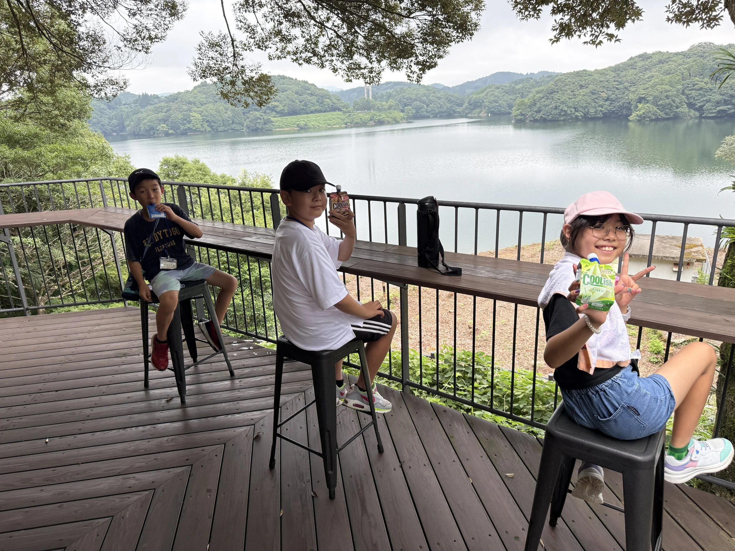 湖の景色を背景に、木製のデッキと手すりのある場所で、3人の子供たちが飲み物を持って座っている写真。左の少年は飲み物を飲んでいる。中央の少年はカップを持ち、右の少女は笑顔でピースサインをしている。