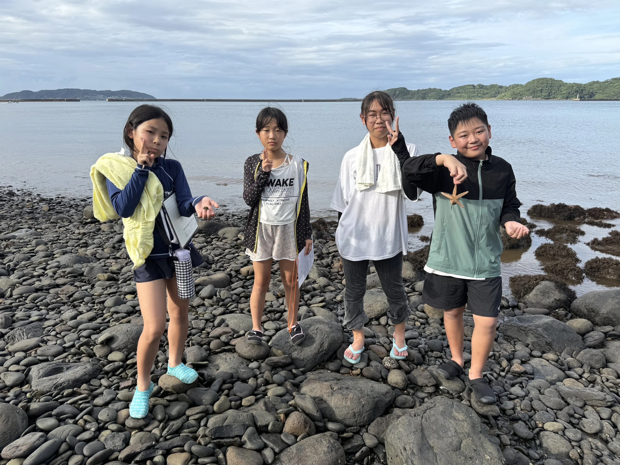 浜辺にいる4人の子供たち。岩や石が散らばっており、海と山々が背景に見える。