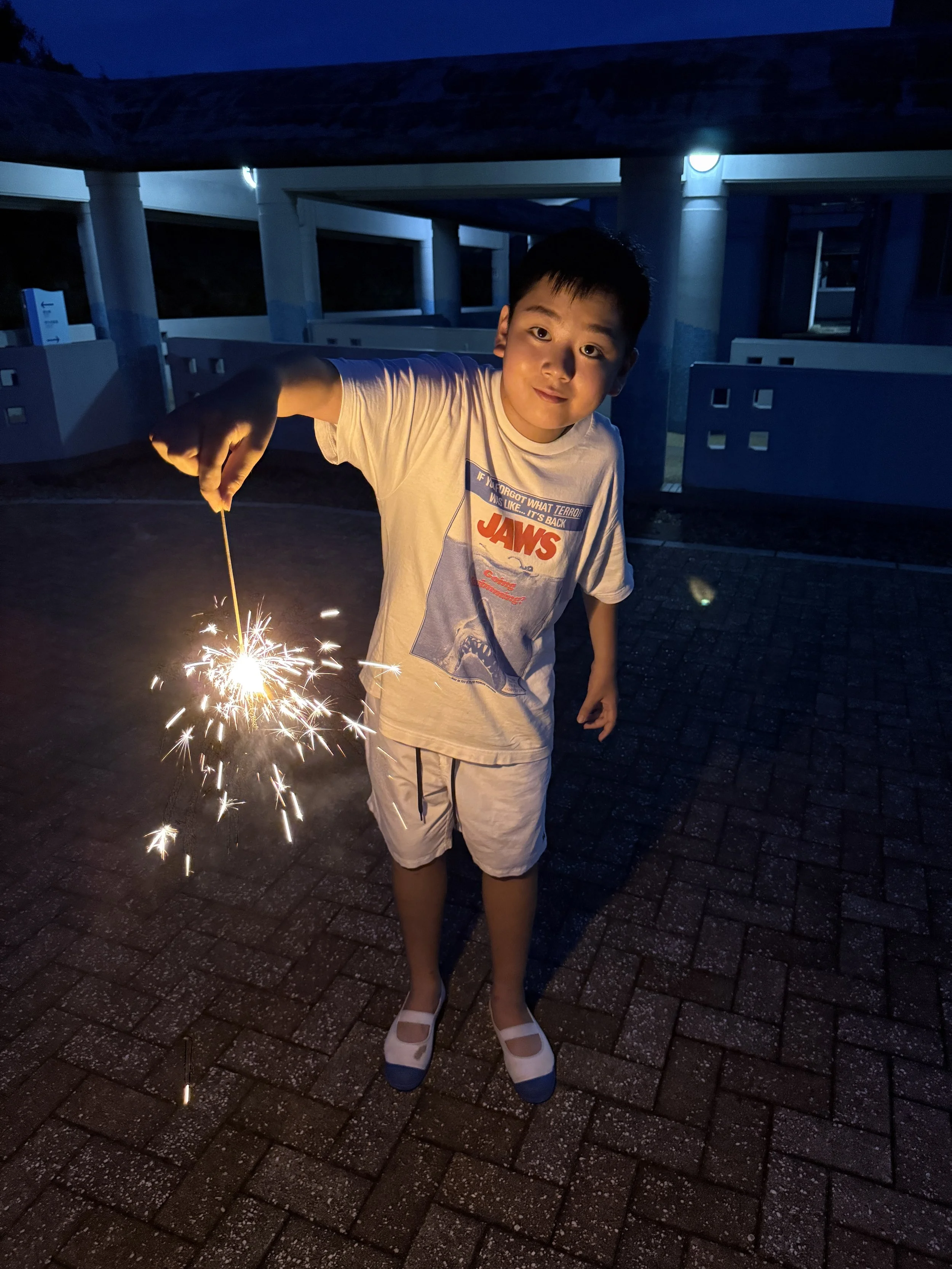 夜に少年が花火を持ち、笑顔で写真を撮っている。少年は白いTシャツと白いショーツ、白と青の靴を履いている。背景は建物と夜空。