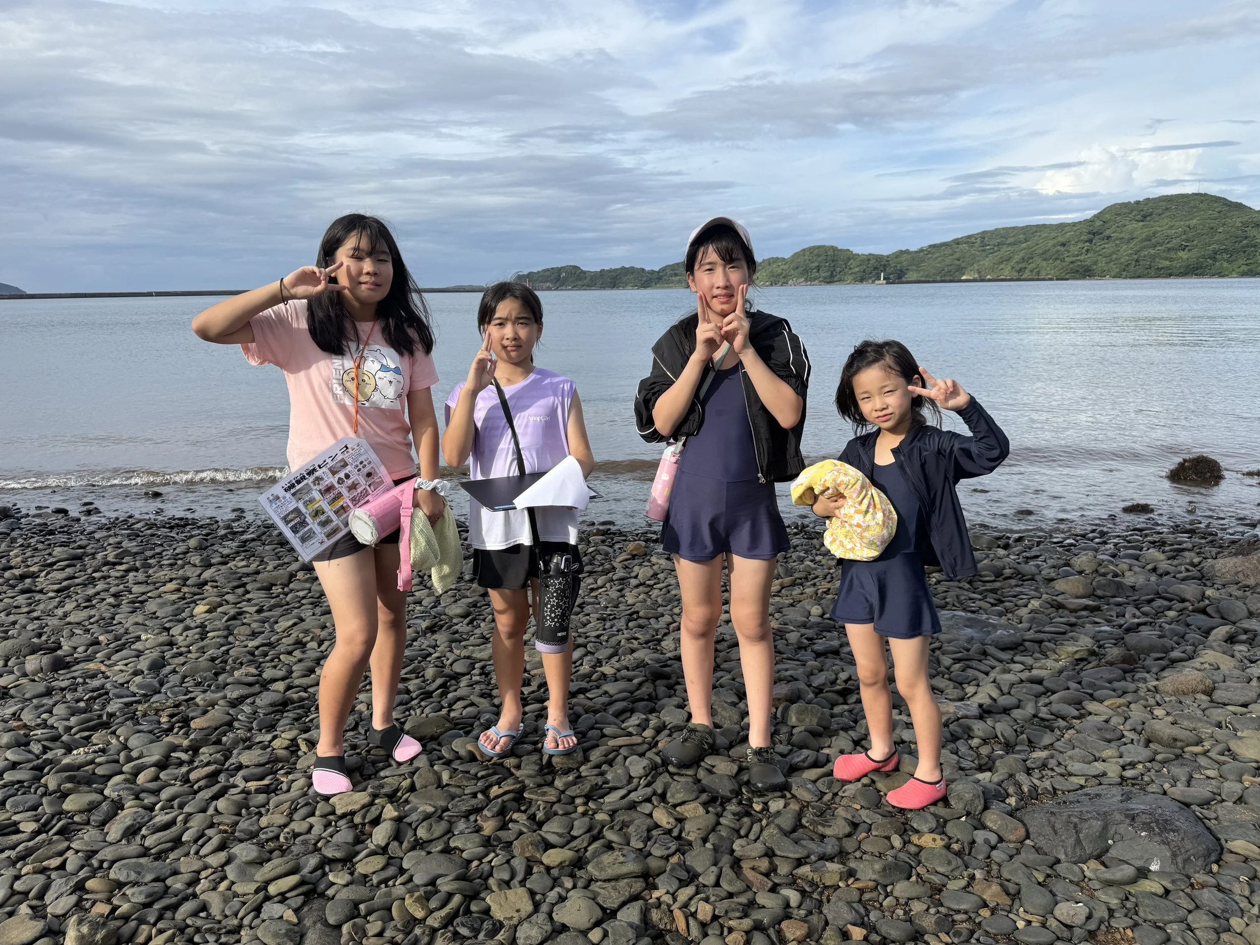 海辺で4人の子供たちがポーズをとっている写真。背後には海と山が見える晴れた日晴天の風景。子供たちはカジュアルな服装とサンダルを履いている。