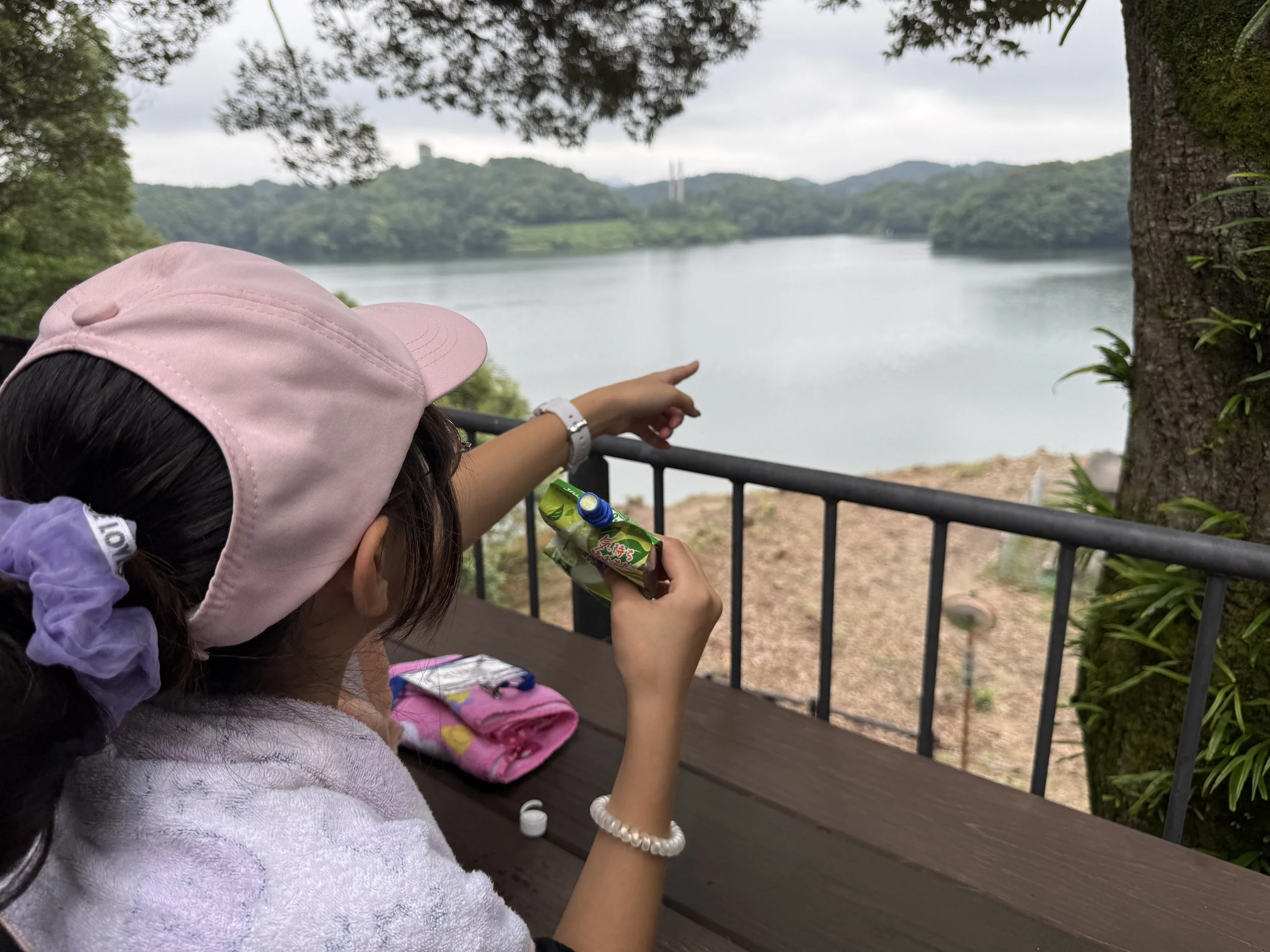 湖の景色を眺めながら座っている女の子が、飲み物を手に持ち、指を湖の方向に向けている。背後に木と山々が見える