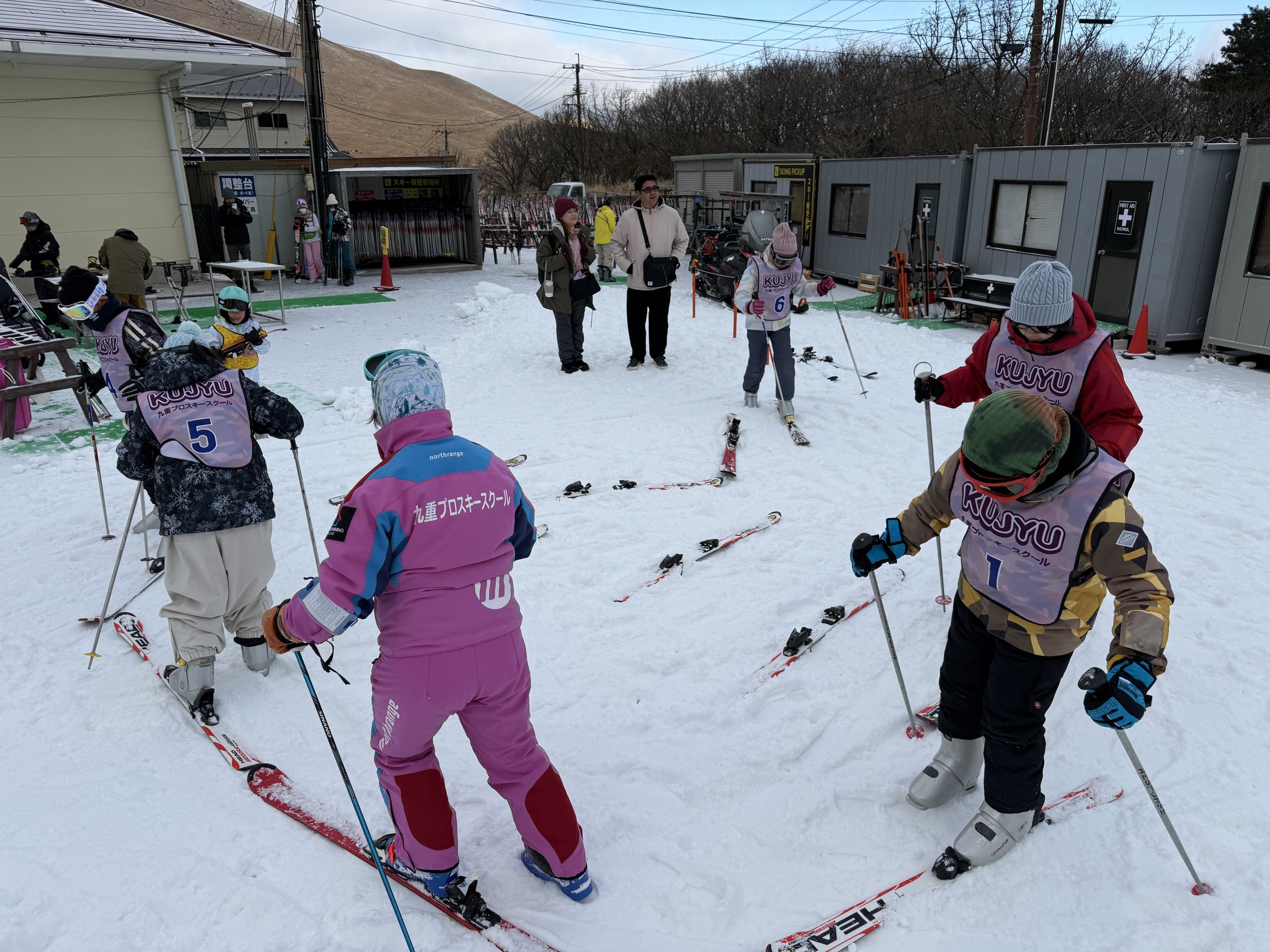 雪の上でスキーを履いている子供たちと大人たち。指導者や参加者が集まるスキー教室の外観風景。背景に建物や人々、山や木々が見える。
