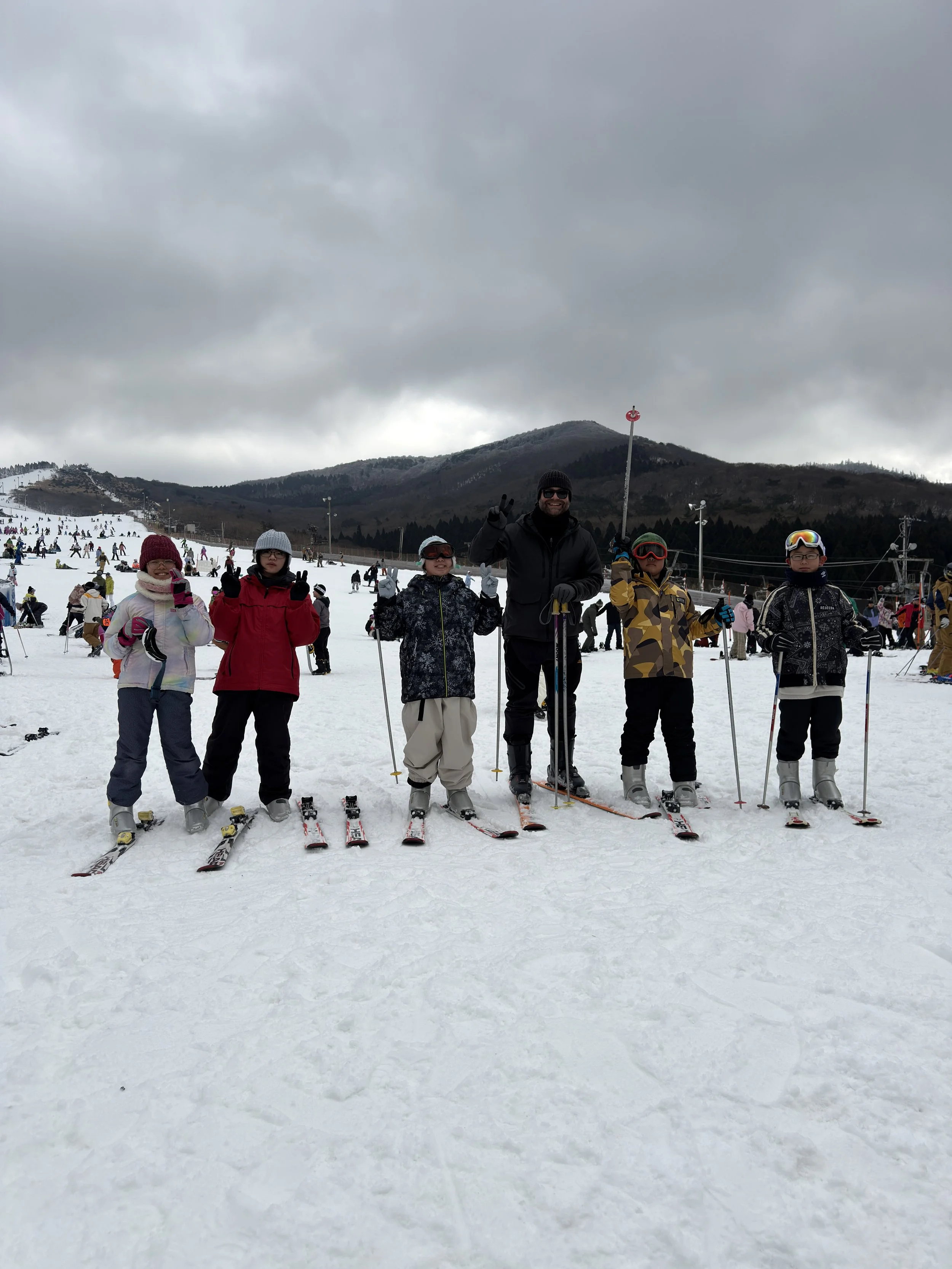 雪山でスキーをしているグループの写真。5人の成人と子供たちが笑顔でポーズをとっている。背景に他のスキーヤーや雪山が見える。