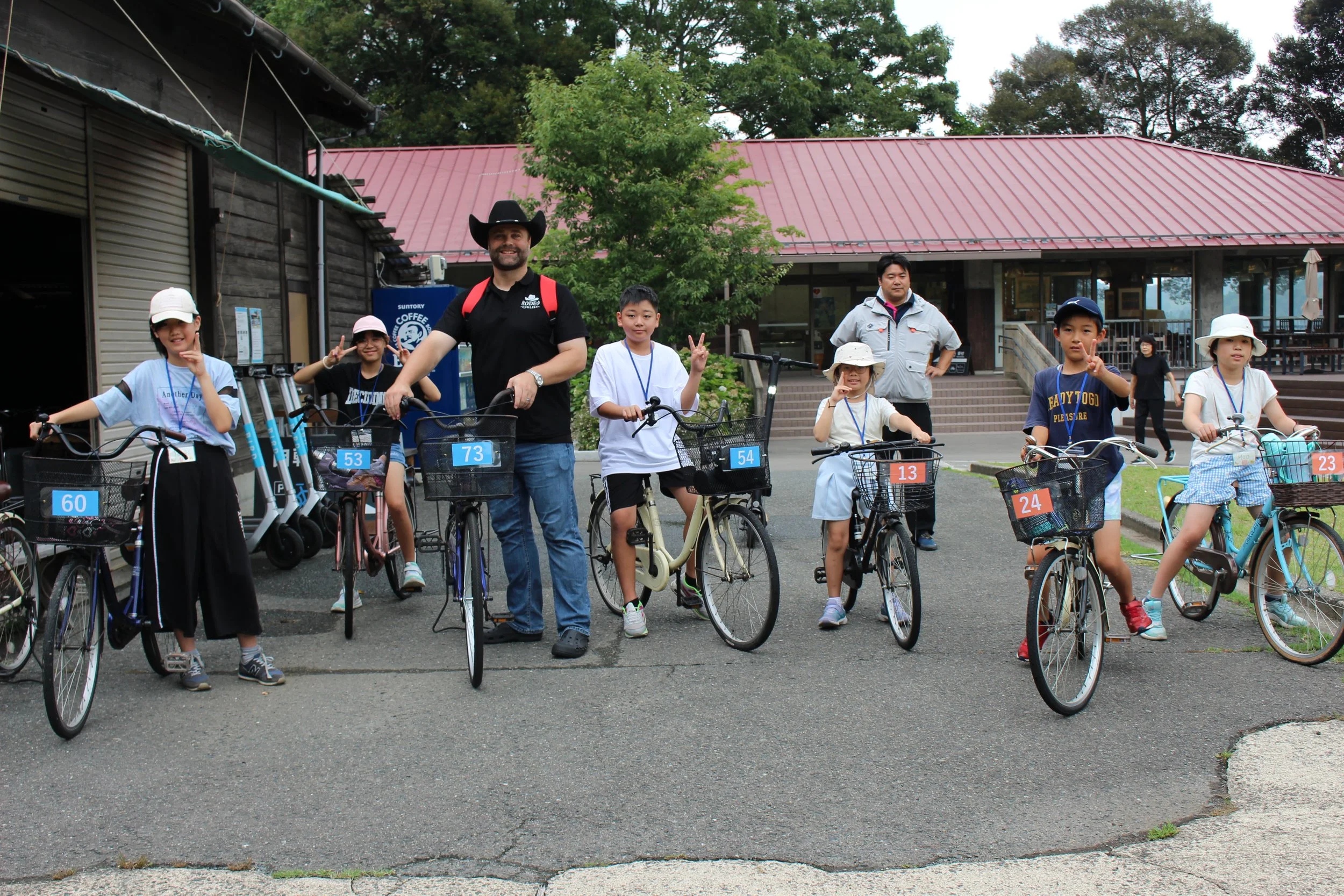 子供たちと大人が自転車に乗っているグループ写真。背景に建物と緑の木々がある。