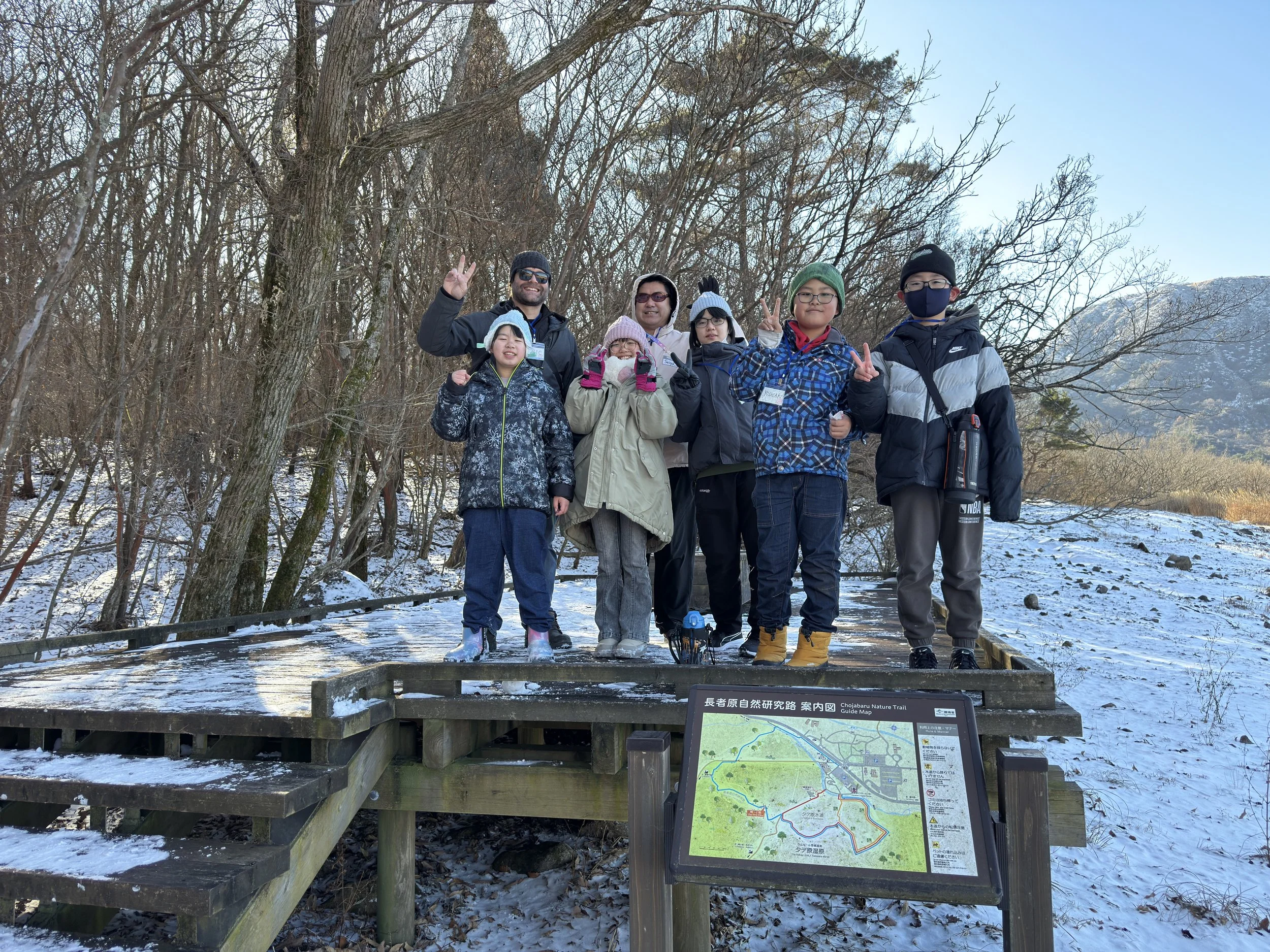 冬の自然の中で観光客のグループが写真を撮っている。地面に雪があり、木々は葉を落としている。グループは多様な年代と性別で、寒冷地の服装をしている。背景には山が見える。