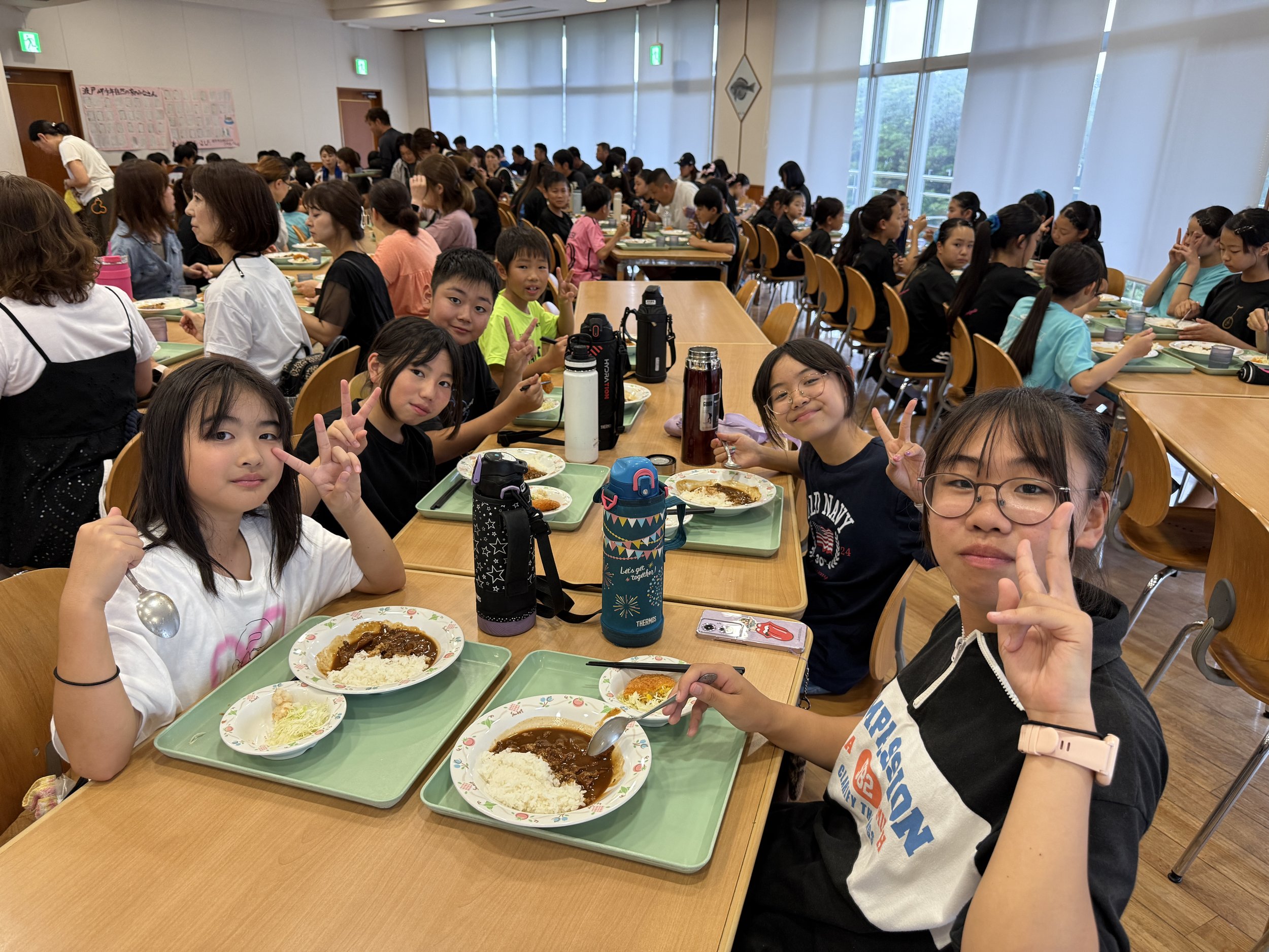 学校の食堂で机に座る子供たちと大人たちがみんな食事をしている。子供たちは手を振ったりピースサインをしている。