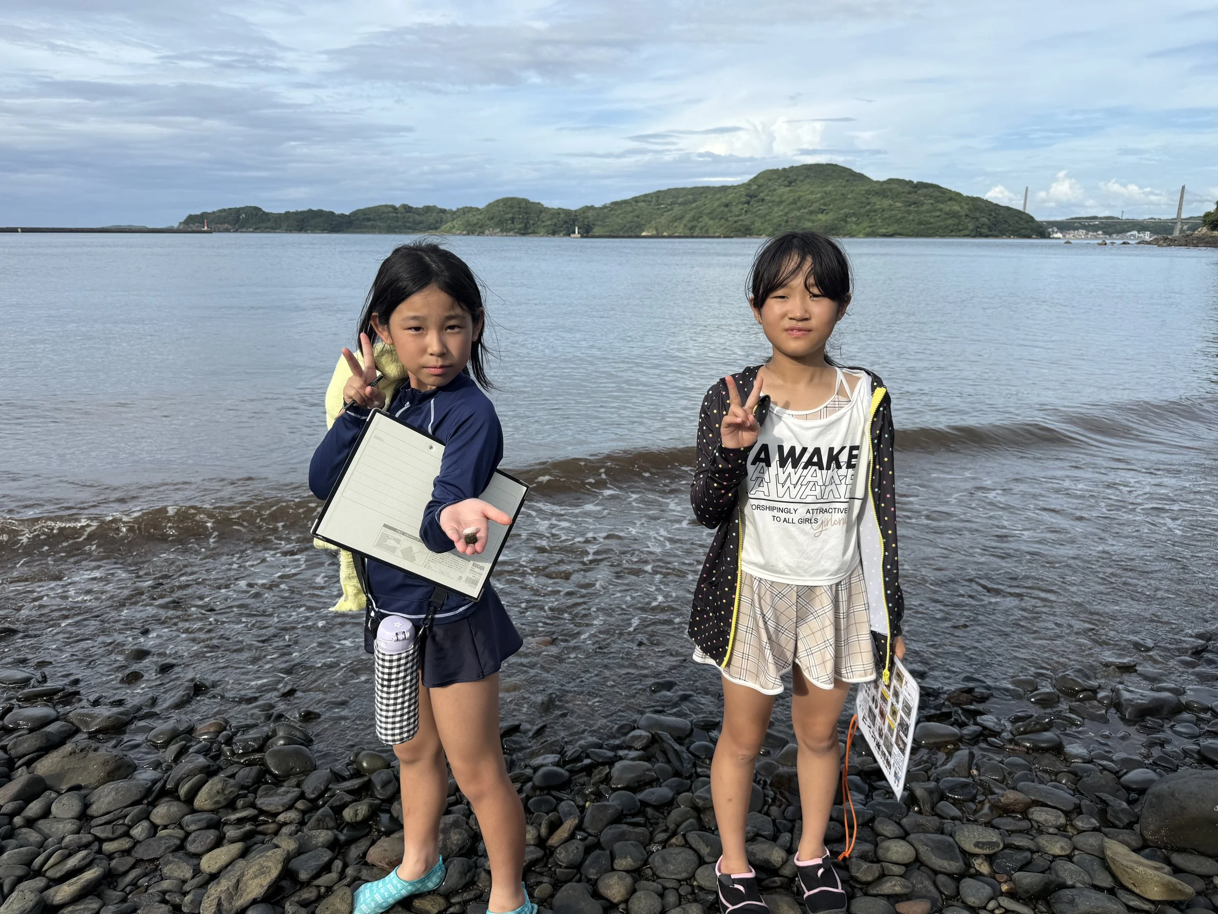 海辺に立つ二人の女の子。左の女の子は中指に海の貝殻を持ち、ノートを持っている。右の女の子はVサインをしている。背景には水面と緑の丘が見える。