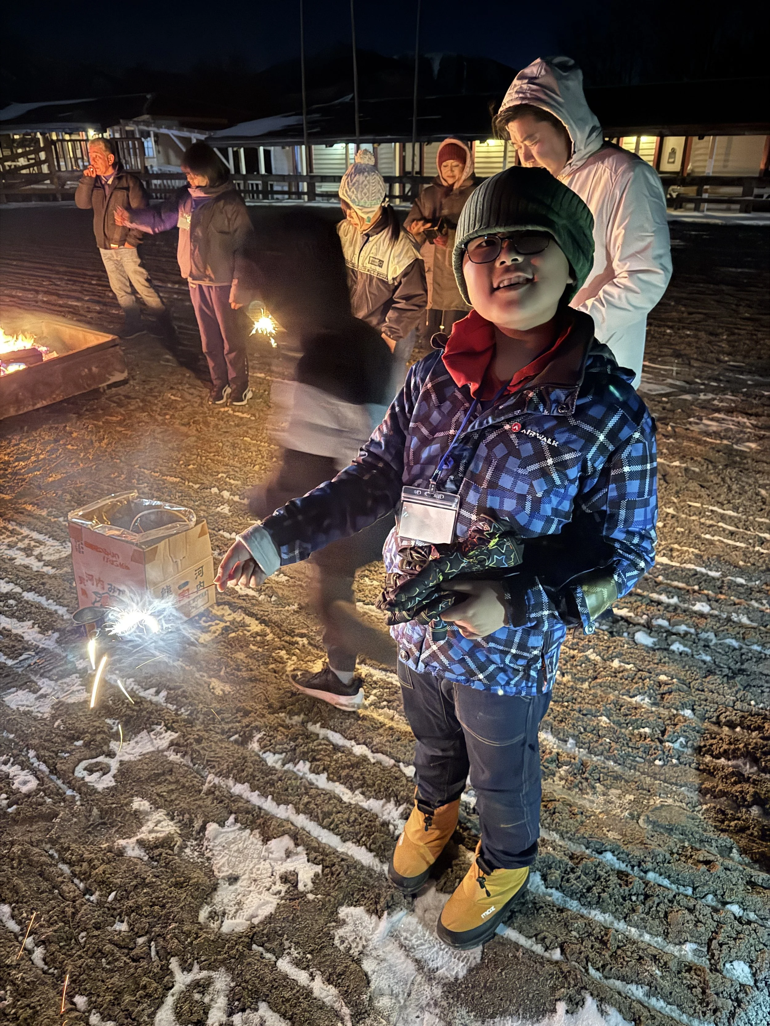 子供と大人たちが夜に屋外で花火を楽しむ光景。少年が花火を持っている。周囲に雪があり、寒そうな服装をしている。