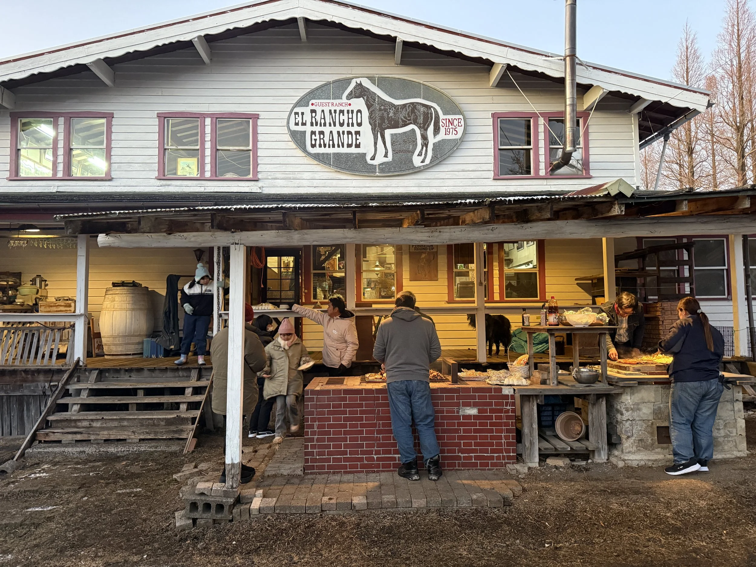 木造のレストランの外観で、看板に馬のイラストと「EL BANCHO GRANDE」などの文字が書かれている。人々が外で食事をしている様子。夕方の暖かい光が差し込む風景。