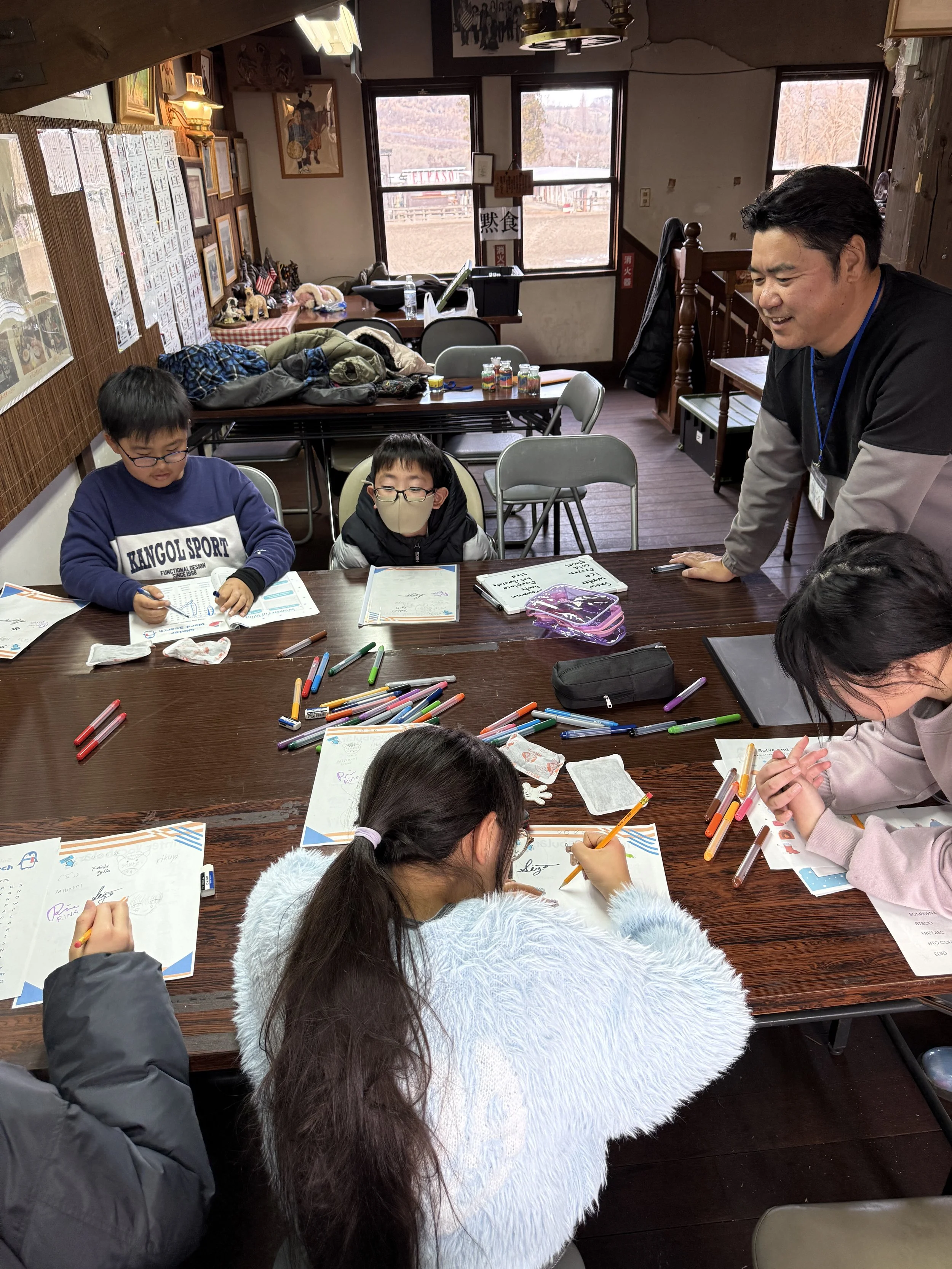 室内のテーブルを囲む子供たちと一人の大人の男性。子供たちは色々な色のペンを使って何かを書いたり描いたりしている。背景に窓や壁に飾られた絵や掲示物が見える。