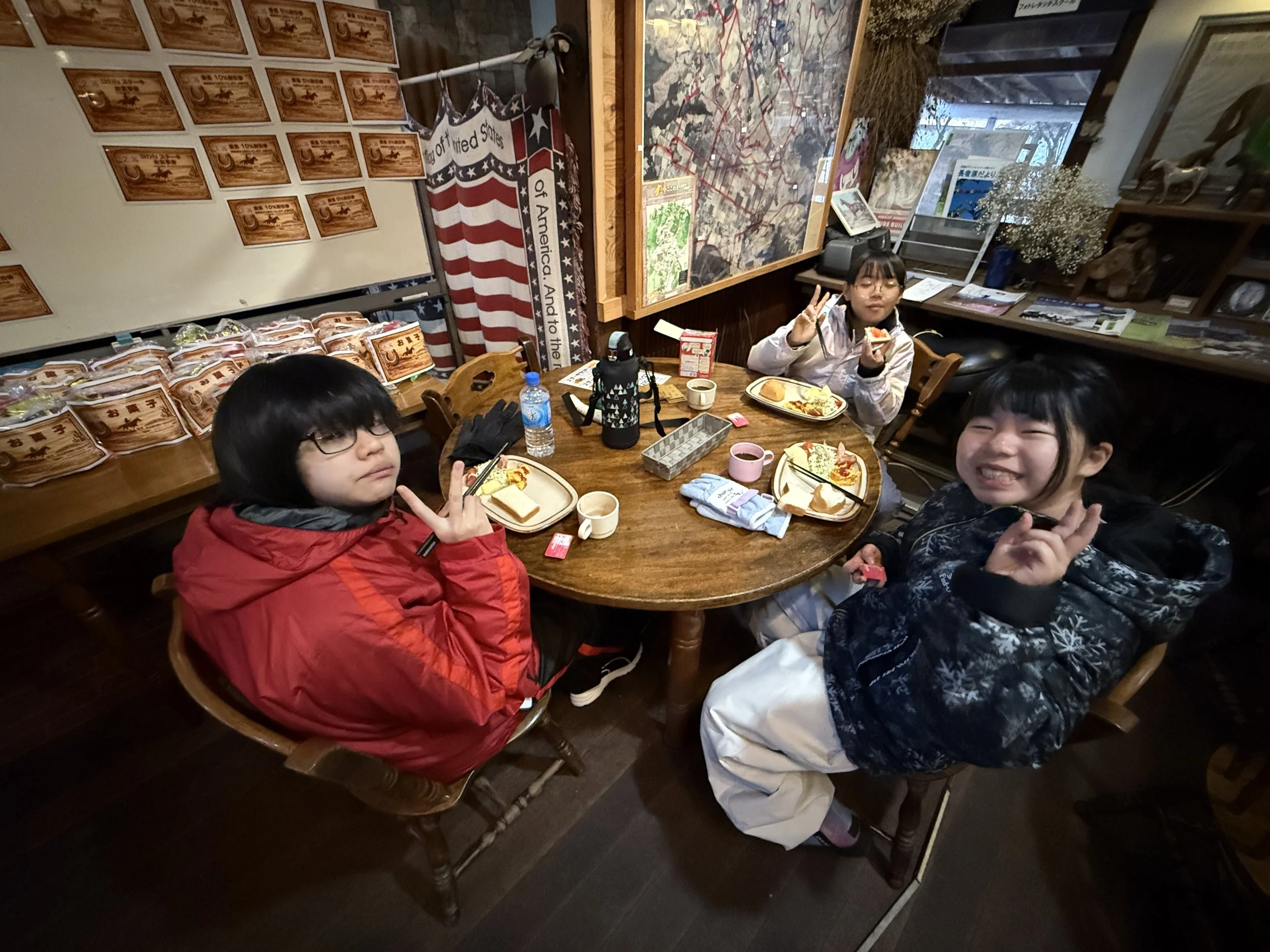 3人の子供がテーブルで食事をしている写真。背景にはアメリカ国旗の装飾や、地図やポスターが貼られている。