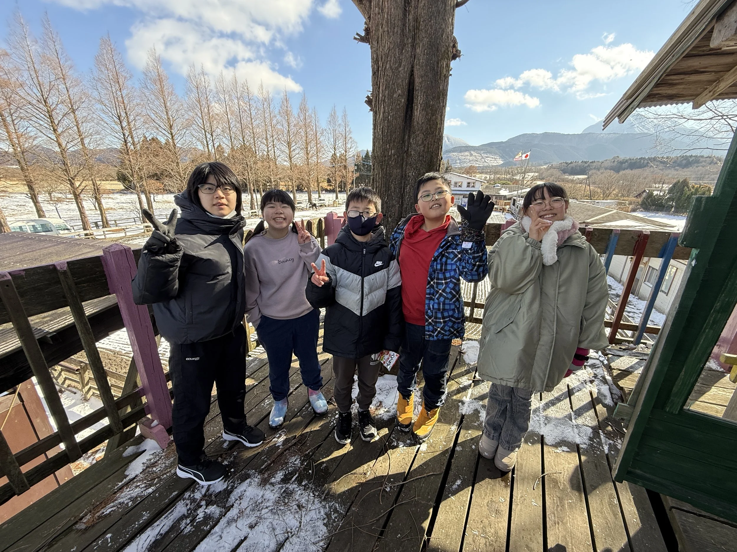 冬の屋外で六人の子供たちが笑顔でポーズをとっている写真。背景に山と日本の旗や木々が見える。