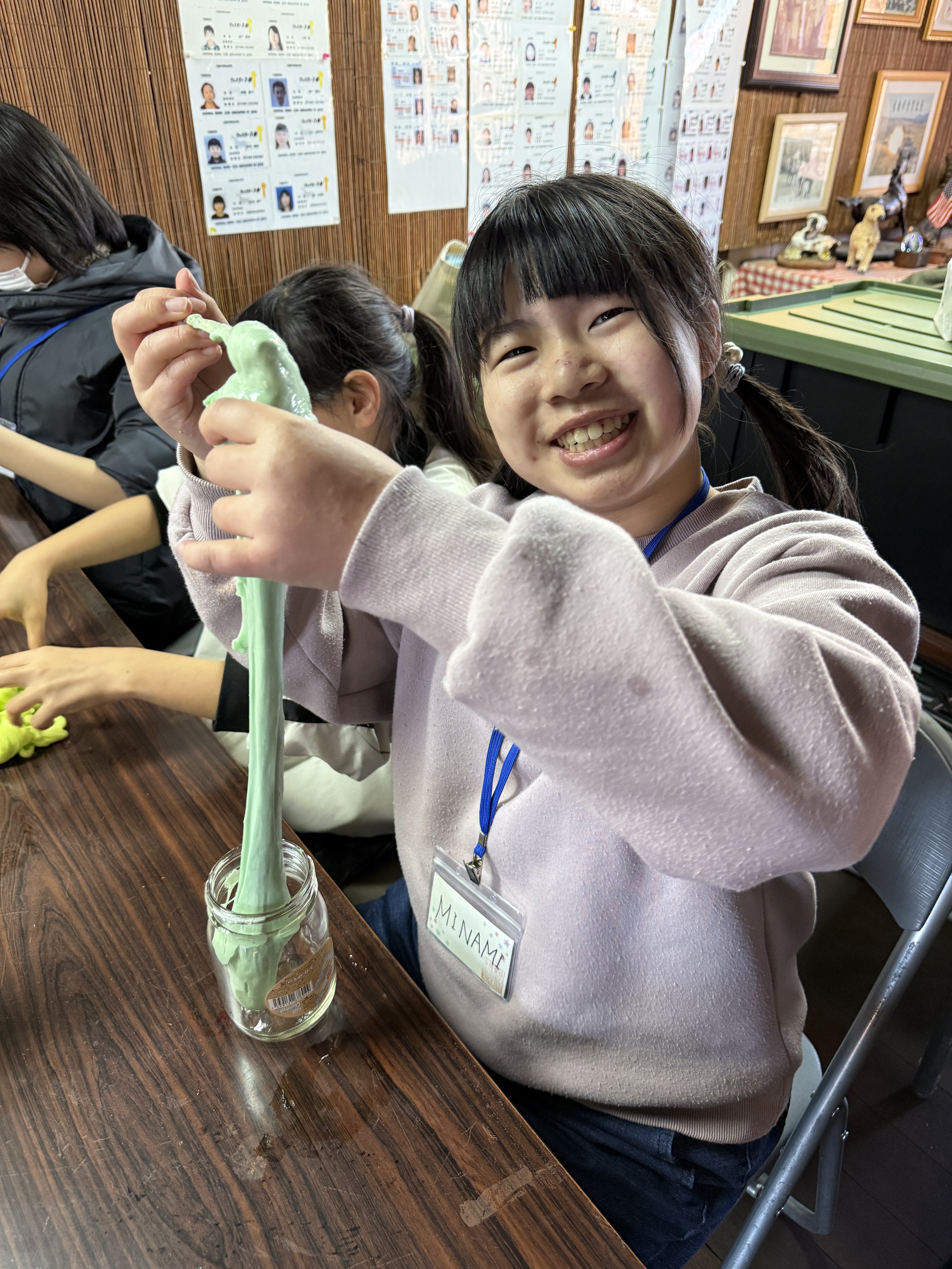笑顔の女の子が緑色の粘土を瓶に伸ばしている。背景には壁に貼られた資料や装飾が見える。