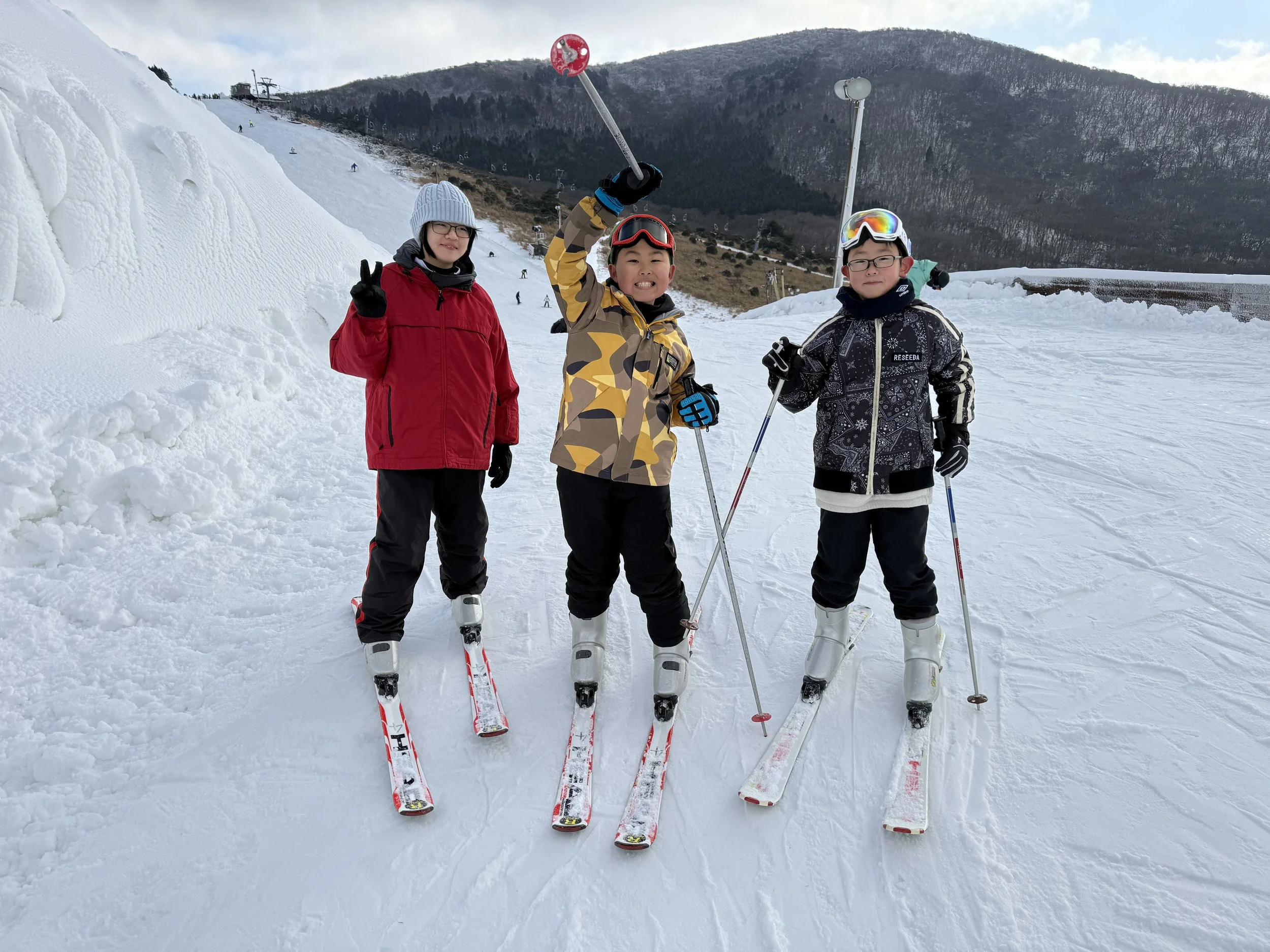 雪山でスキーをする3人の子供たちがポーズをとっている。背景には山と空が広がる。