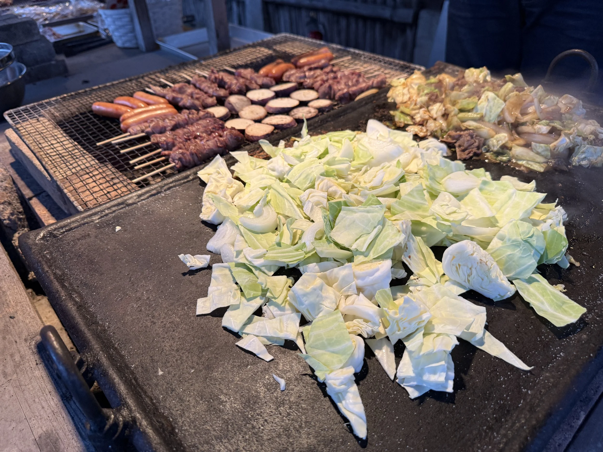 鉄板の上でキャベツと白菜が炒められ、奥では焼きそばやウインナー、肉類がグリルされている。