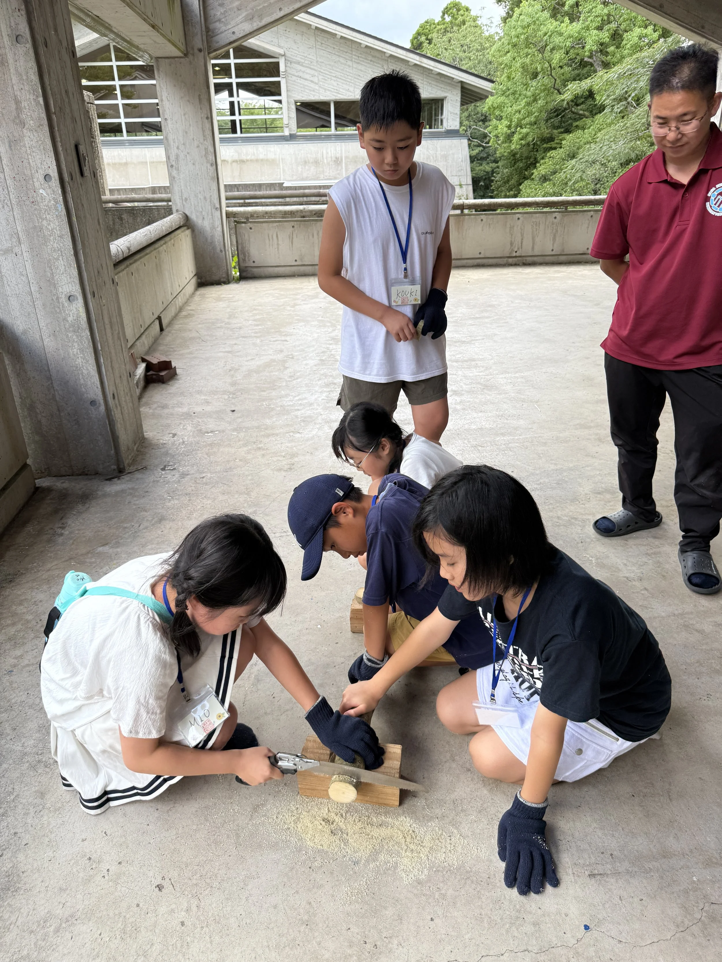 子供たちが木材を使って調理活動をしている様子。2人が木片を叩き割っている。大人とほかの子供たちが見守っている。屋外で行われている活動。