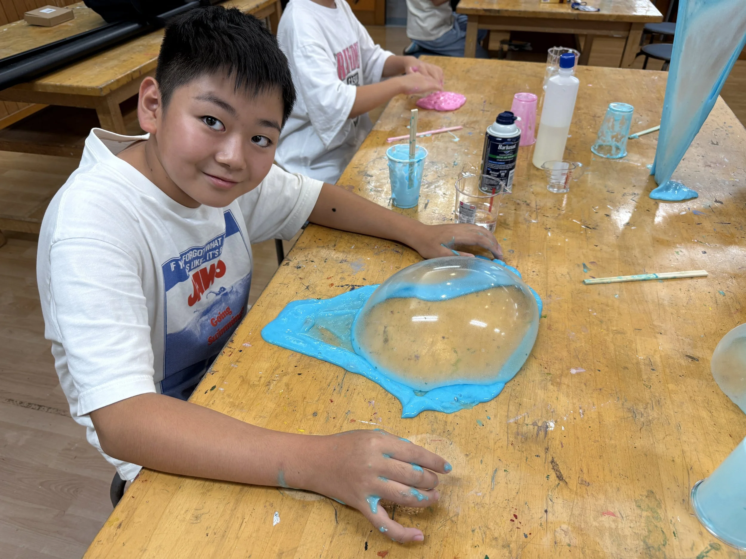 子供が木製のテーブルの上で、青と透明の液体の風船を作っている。背景には他の子供と工作道具が見える。