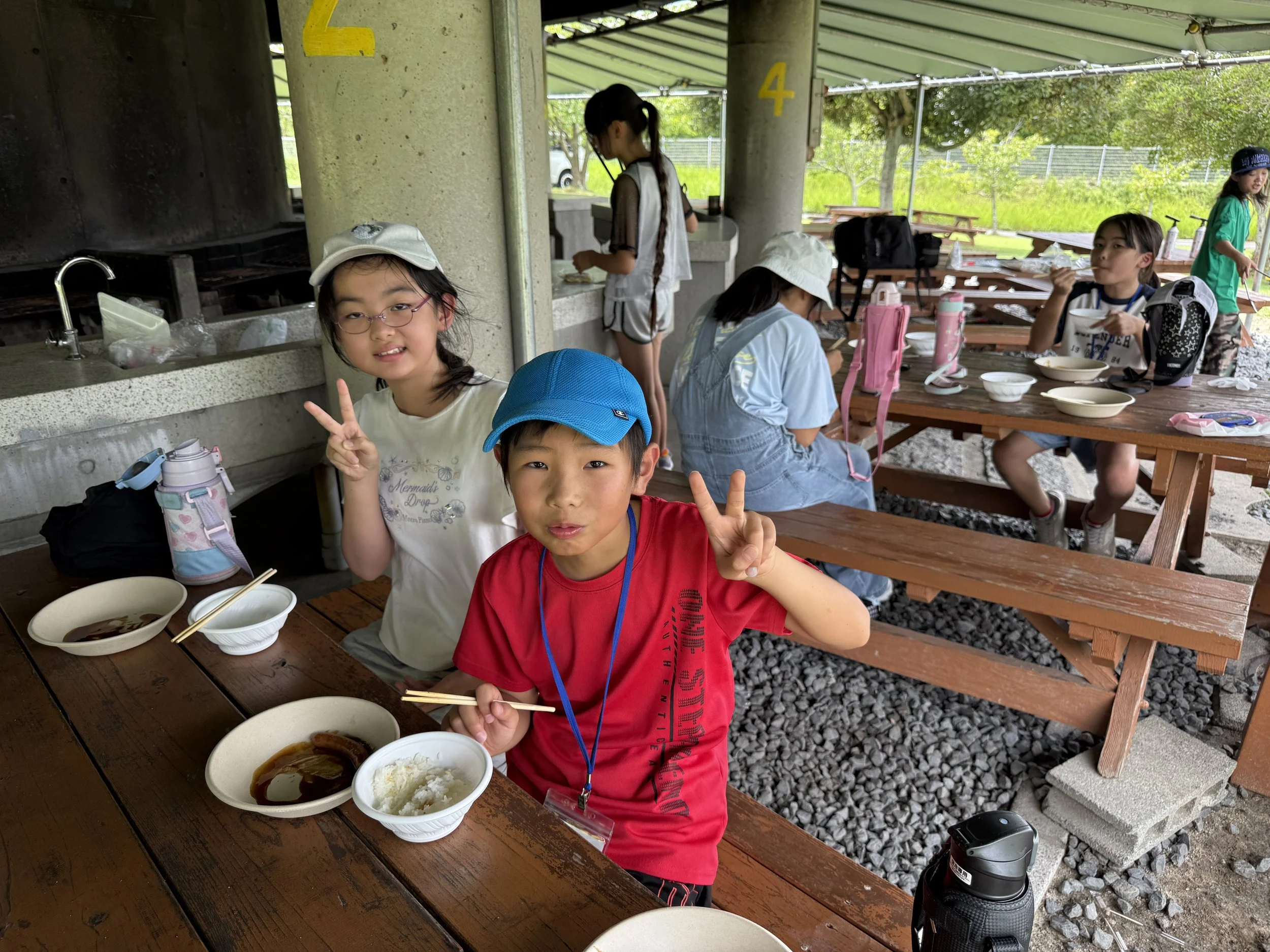 子供たちがバーベキューを楽しんでいる屋外の休憩エリアの写真。子供たちはピースサインをしながら座っており、料理や飲み物がテーブルにあります。背景には緑の木々と屋根が見えます。
