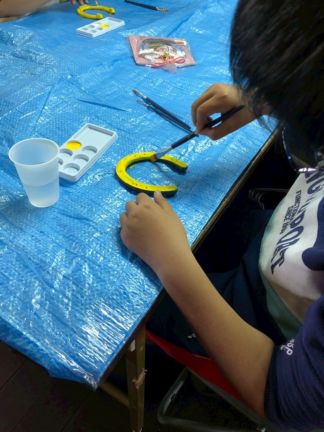 子供が青いテーブルクロスの上で金属製の馬蹄形の飾りをペイントしている様子。