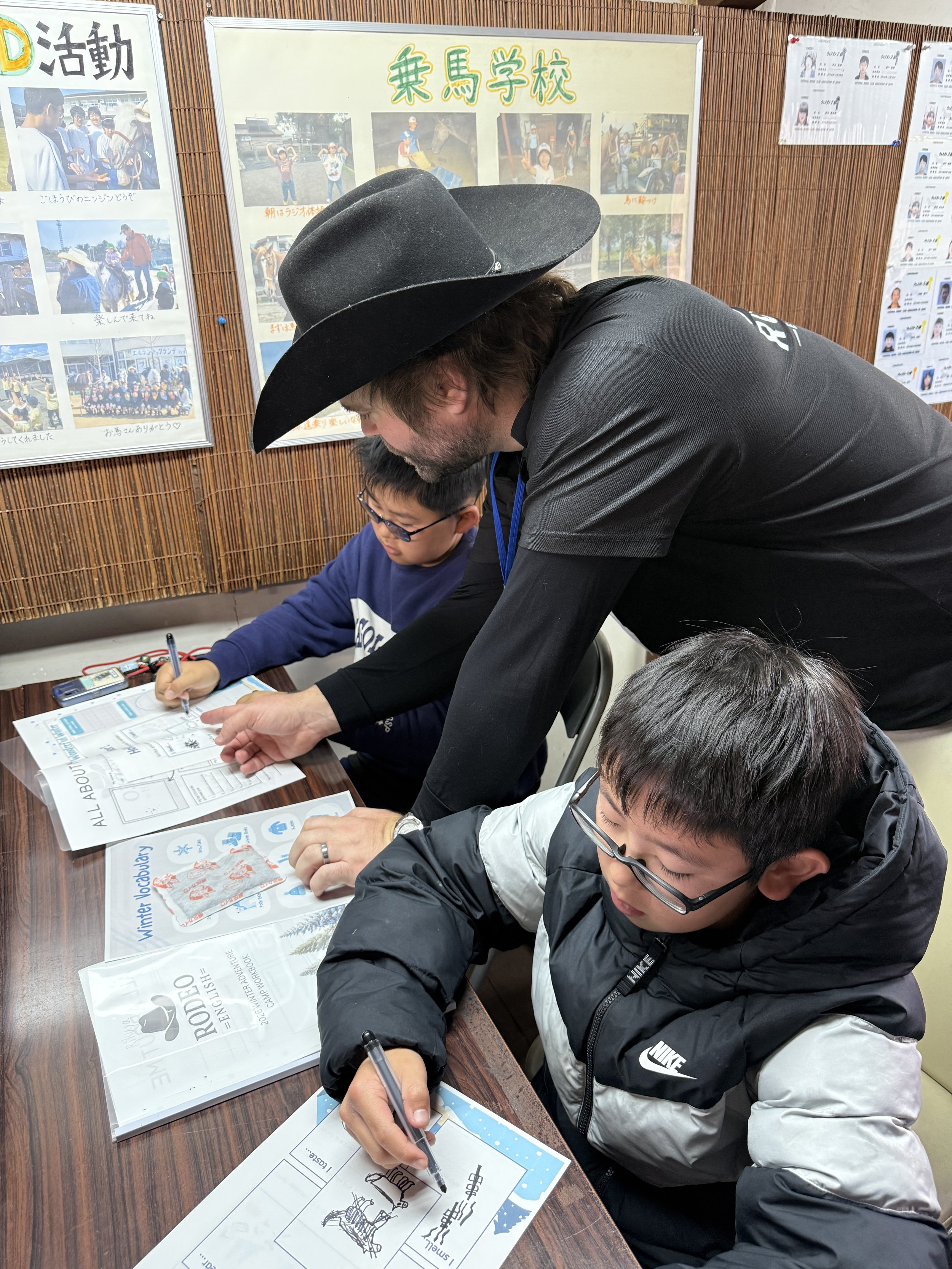 帽子をかぶった親しみやすい男性先生が、二人の子供と一緒に工作や学習に取り組む教室の様子。背景には school's 写真や活動紹介の掲示板が見える。