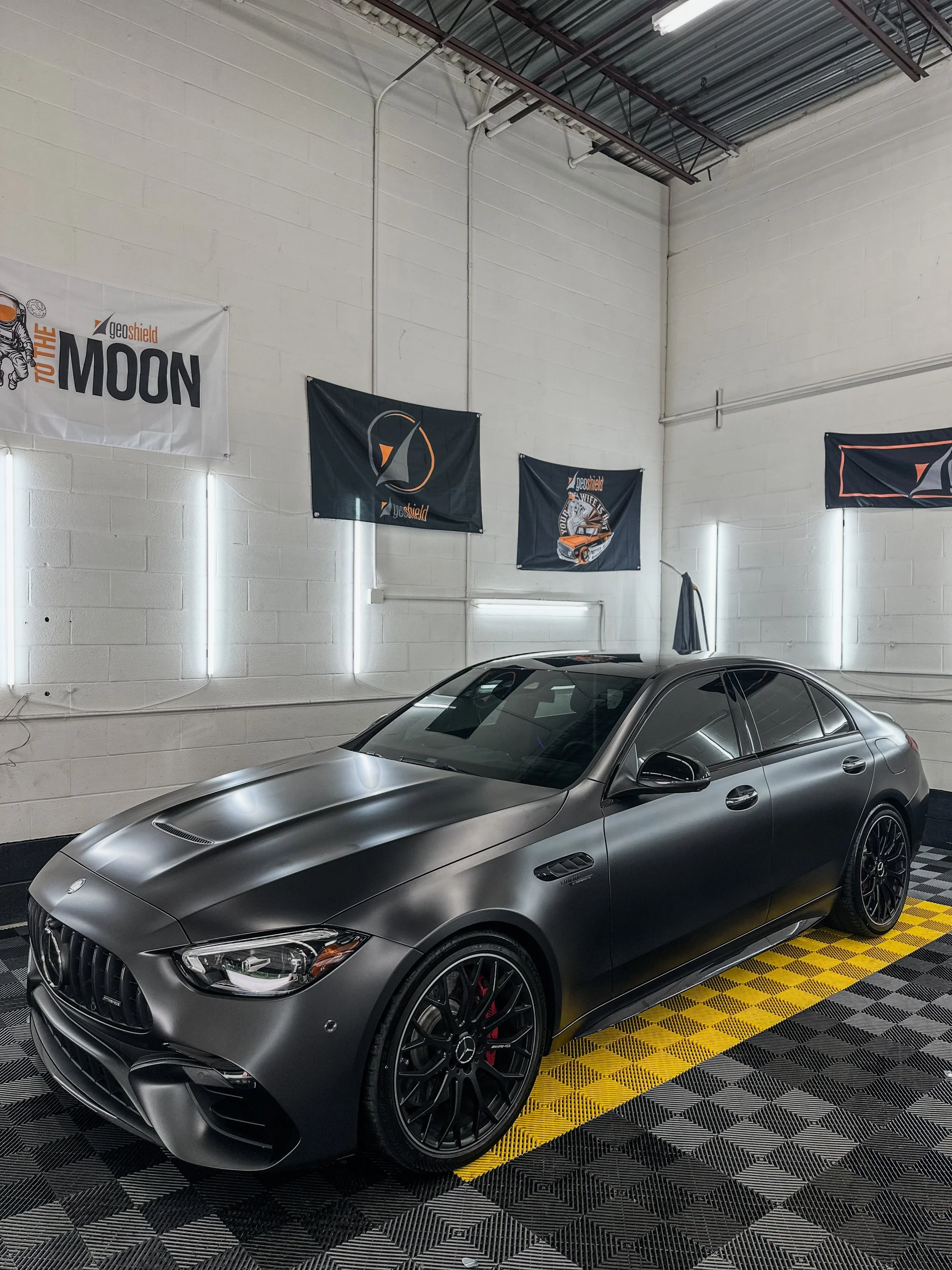 A matte black luxury sedan with tinted windows, black wheels, and red brake calipers inside a well-lit garage or showroom with flags hanging on the walls.