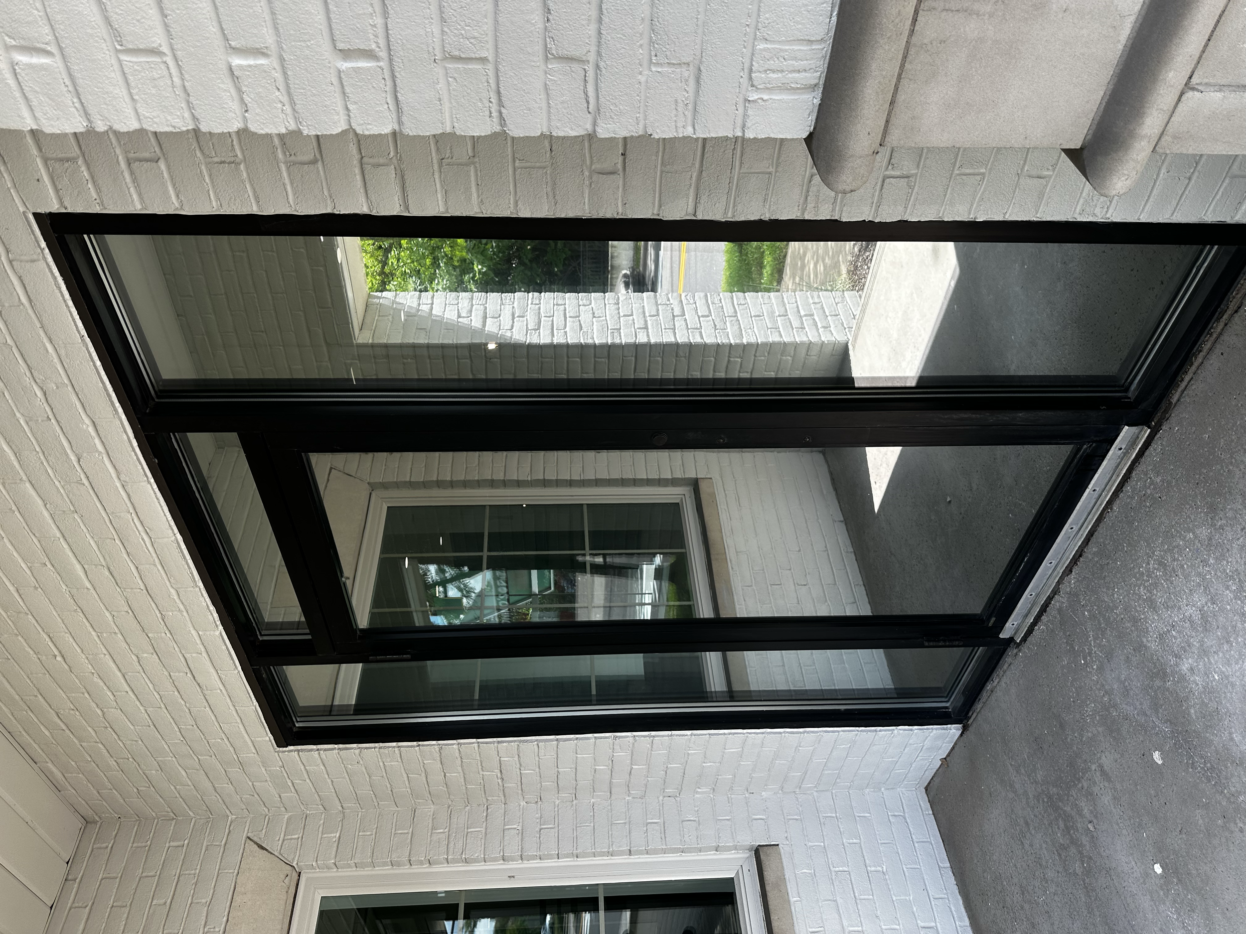 View of a black metal and glass sliding door leading to a small porch area with a concrete floor, white brick walls, an overhead concrete pipe, and a window reflecting trees outside.