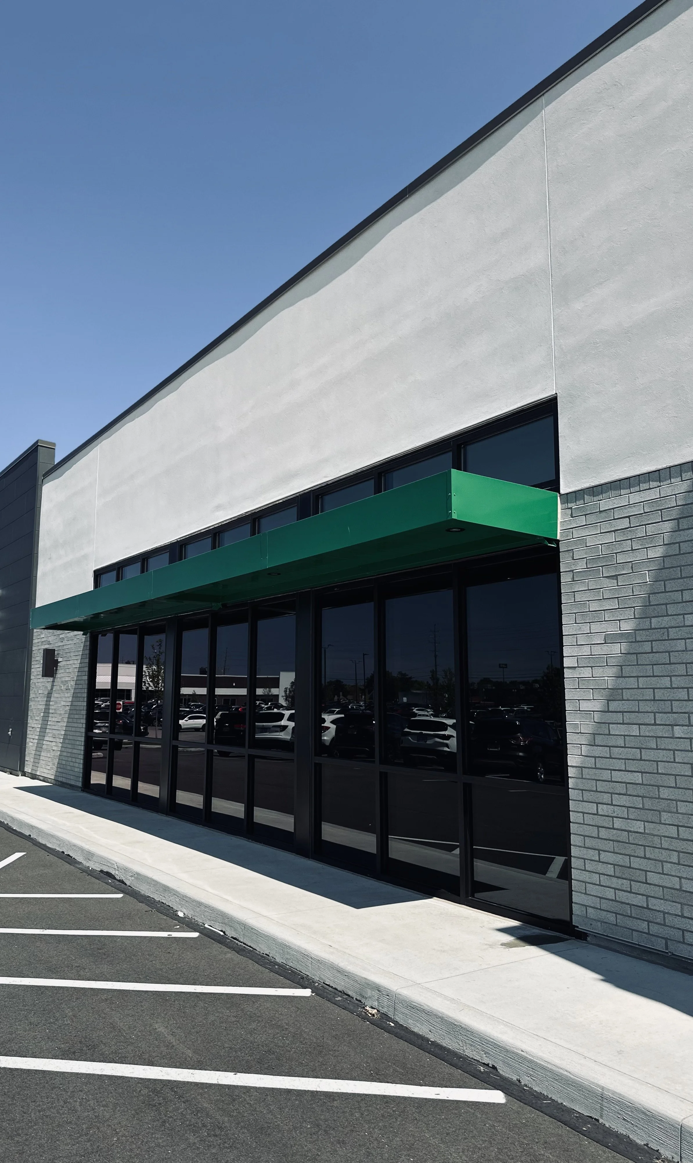 Exterior view of a modern commercial building with large glass tinted windows, a green awning, and a parking lot with multiple cars, under a clear blue sky.