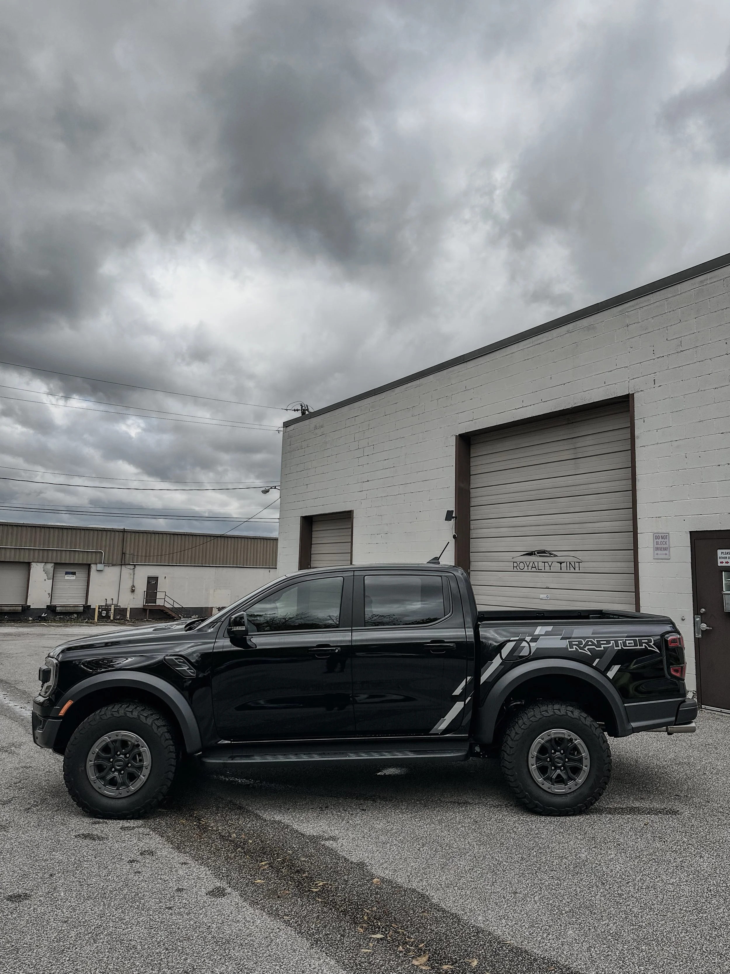 Black pickup truck parked in front of a white industrial building under cloudy sky.