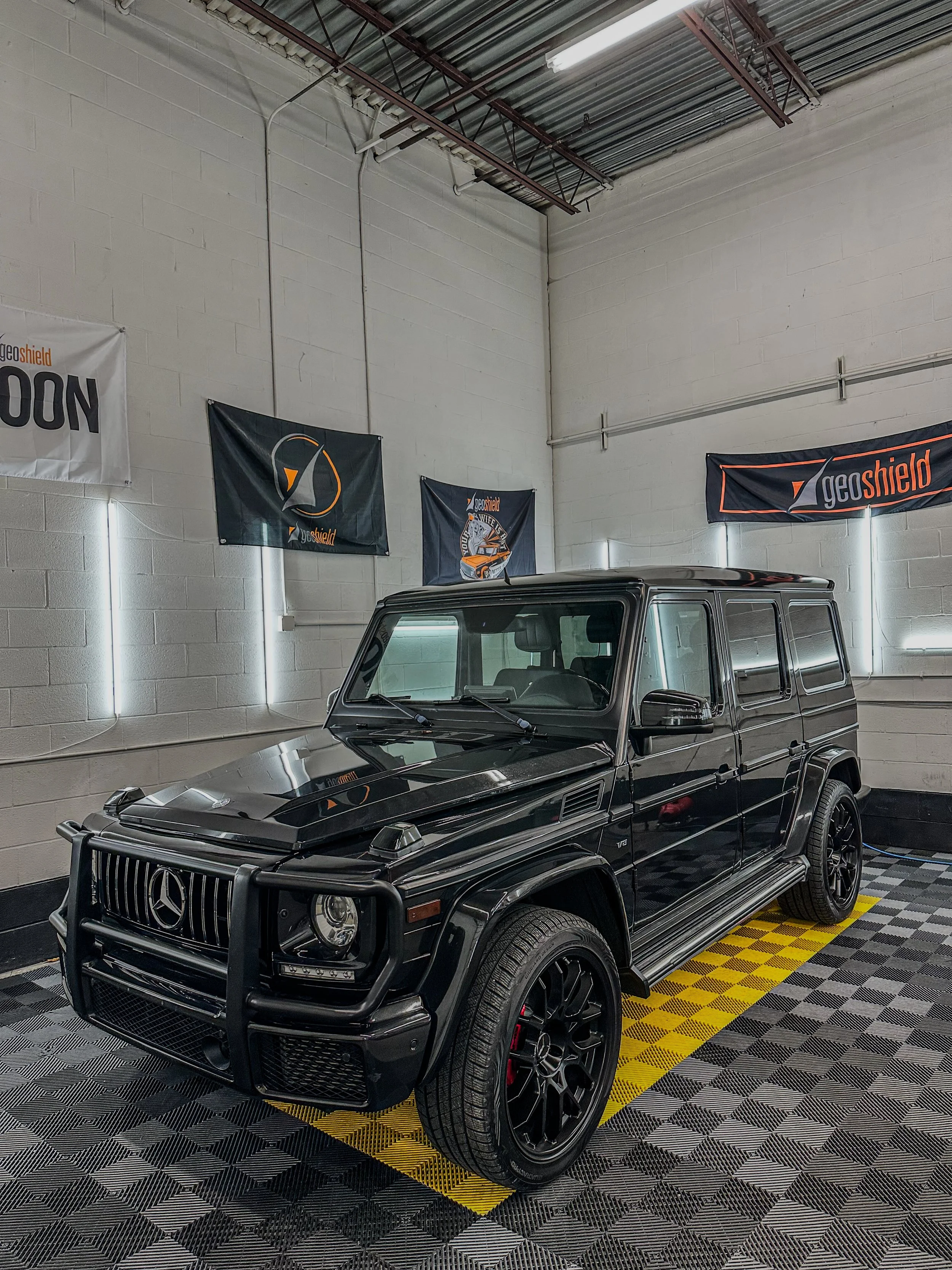 Black Mercedes-Benz G-Class SUV parked indoors on a patterned floor with flags on the wall behind it.