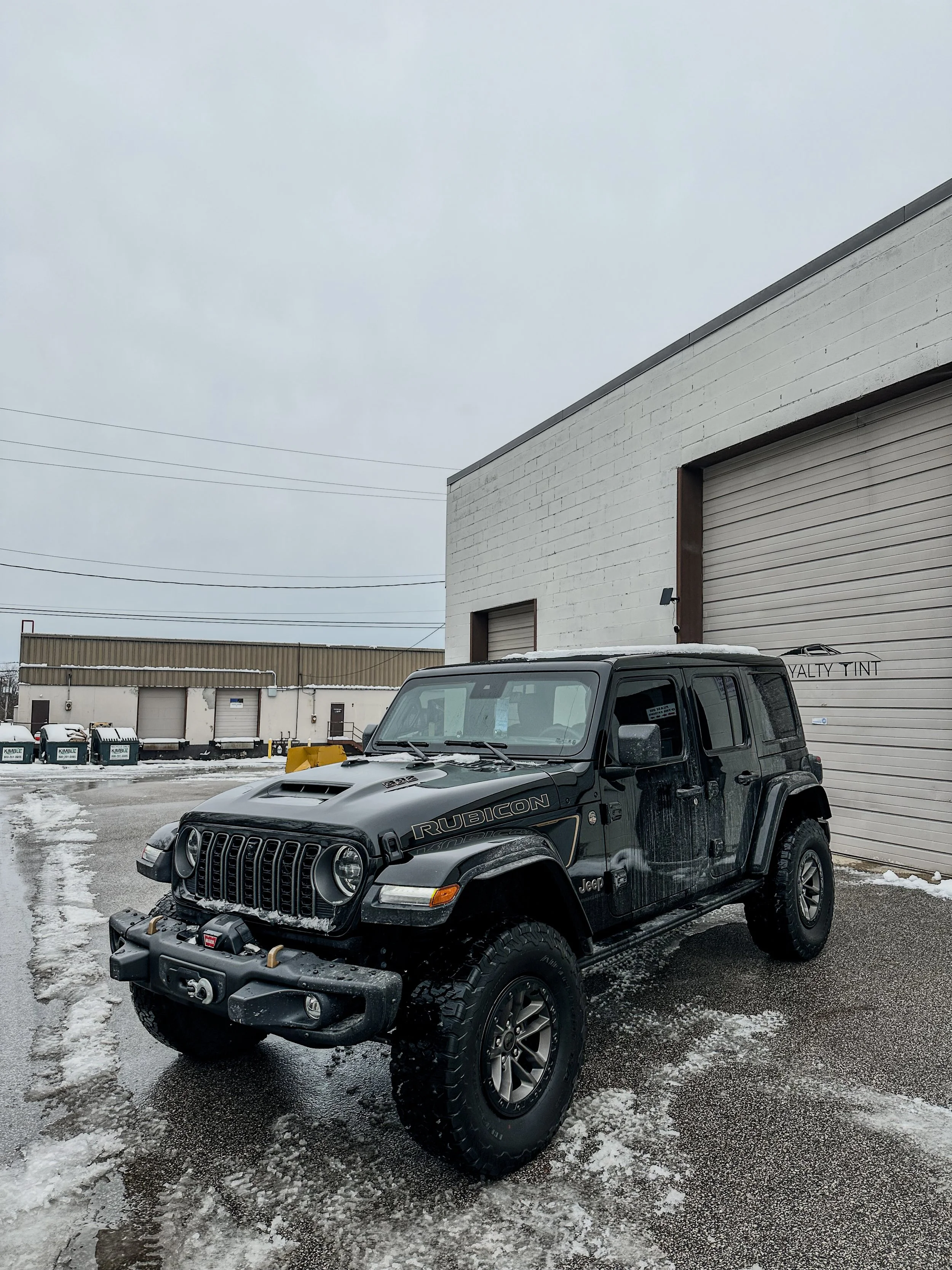 Black Jeep Rubicon parked on wet, snowy ground in front of a white industrial building with closed garage doors.