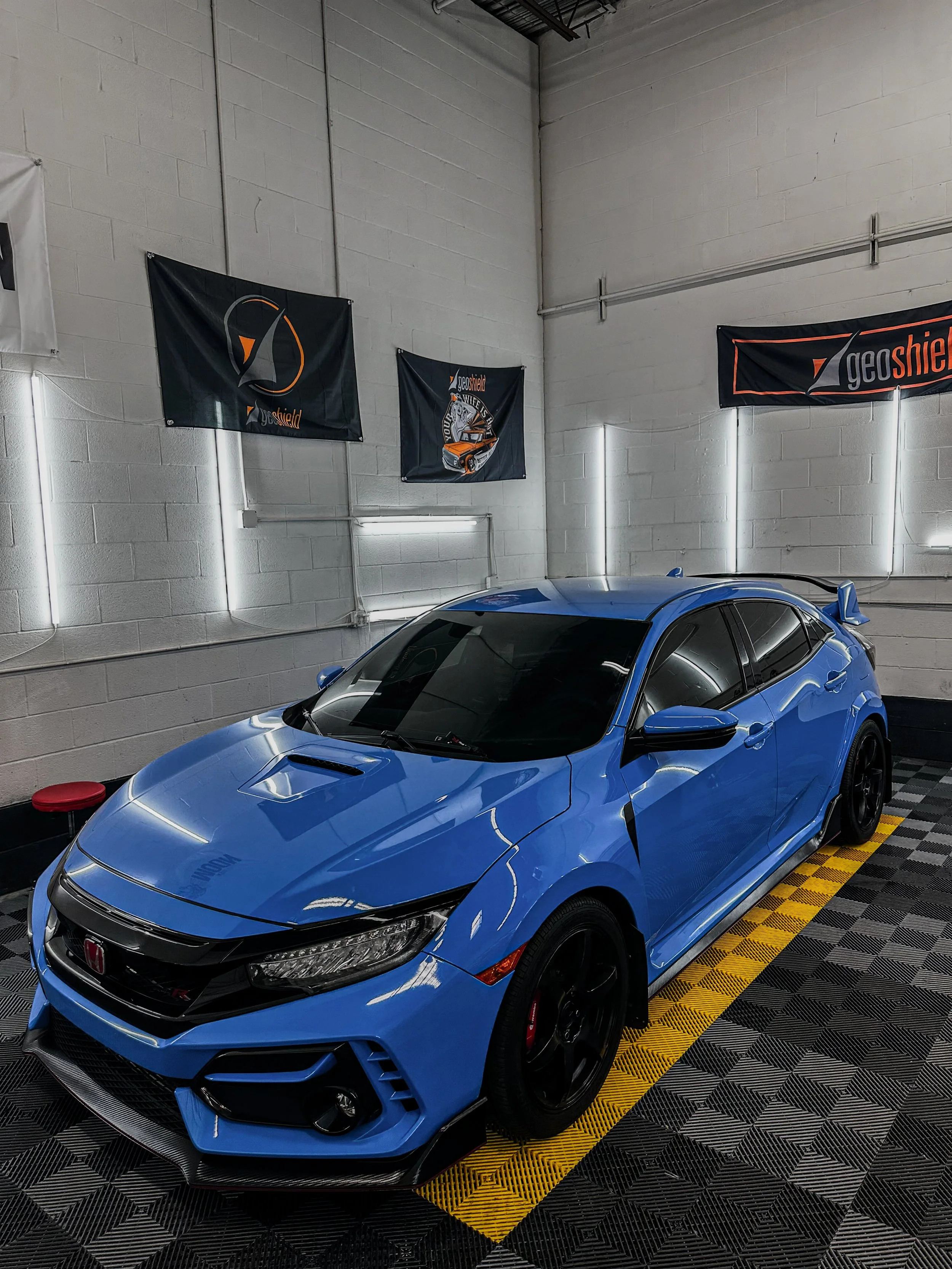 A blue Honda Civic Type R parked indoors on a patterned flooring with black and yellow lines. The background includes white brick walls with banners and racing flags, and a red stool in the left corner.