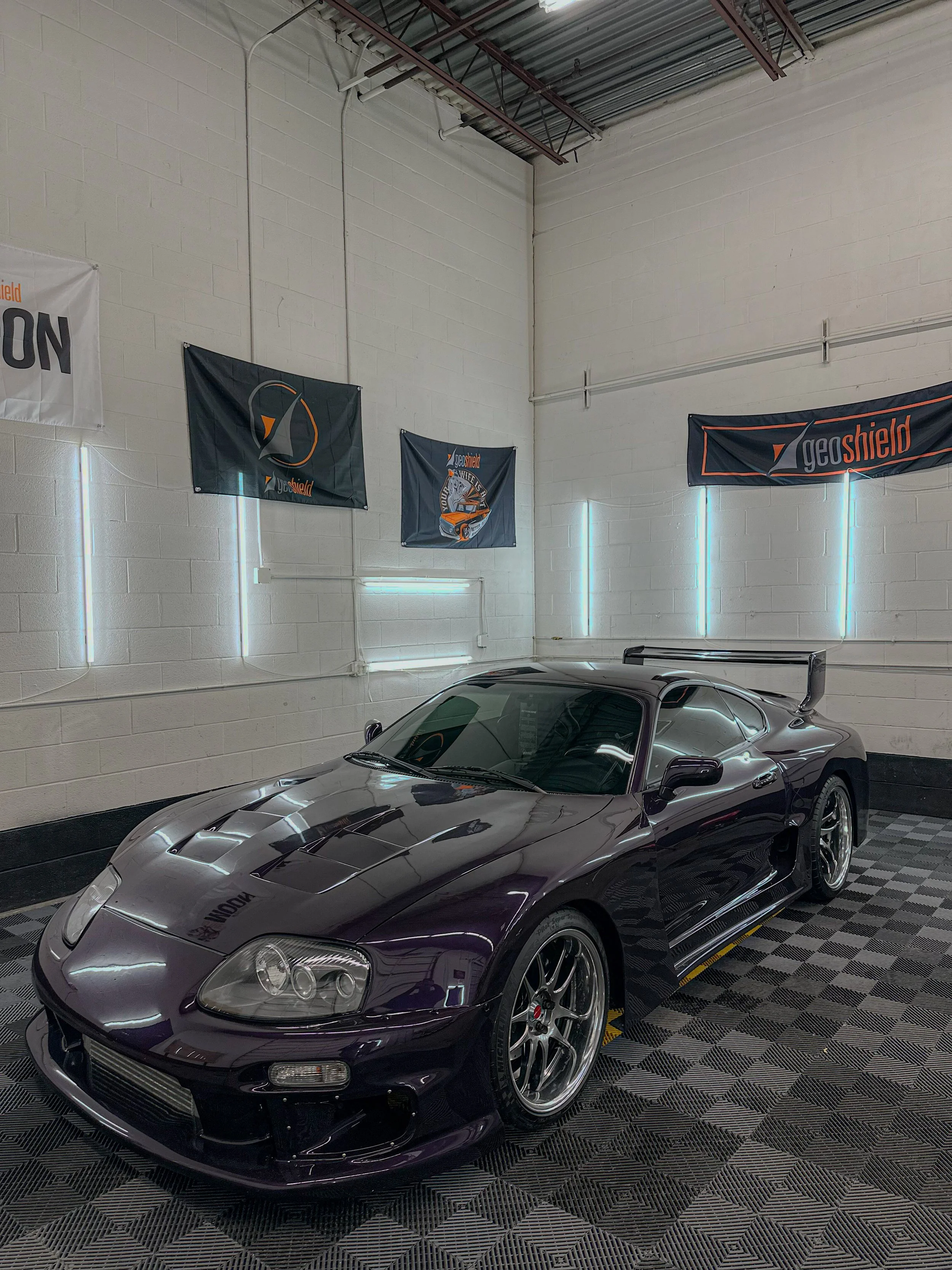 A dark purple Toyota Supra sports car inside a garage or showroom, with flags and neon lights on the wall behind it.