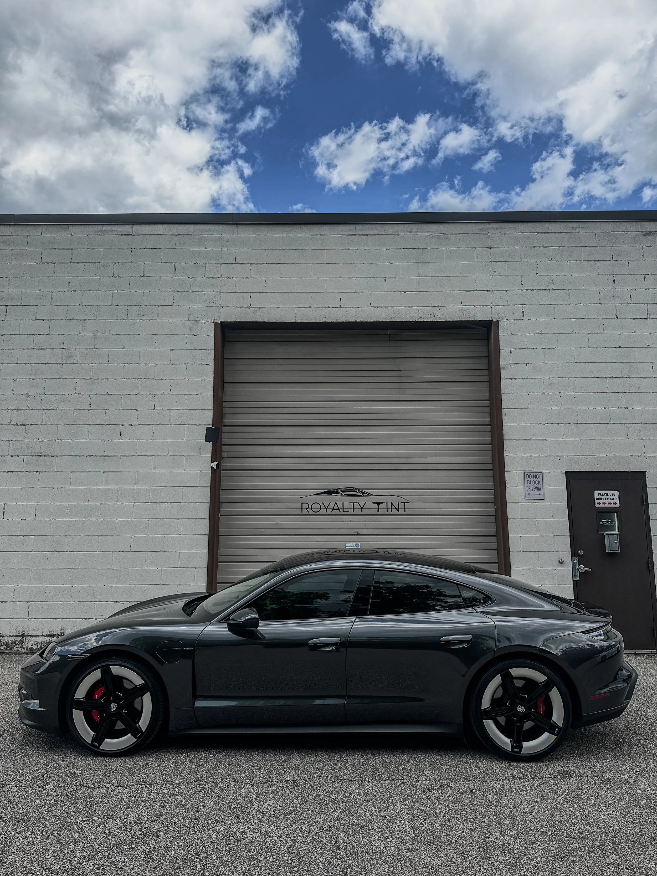A black sports car parked in front of a gray industrial building with a large metal door and a small black door, under a partly cloudy sky.