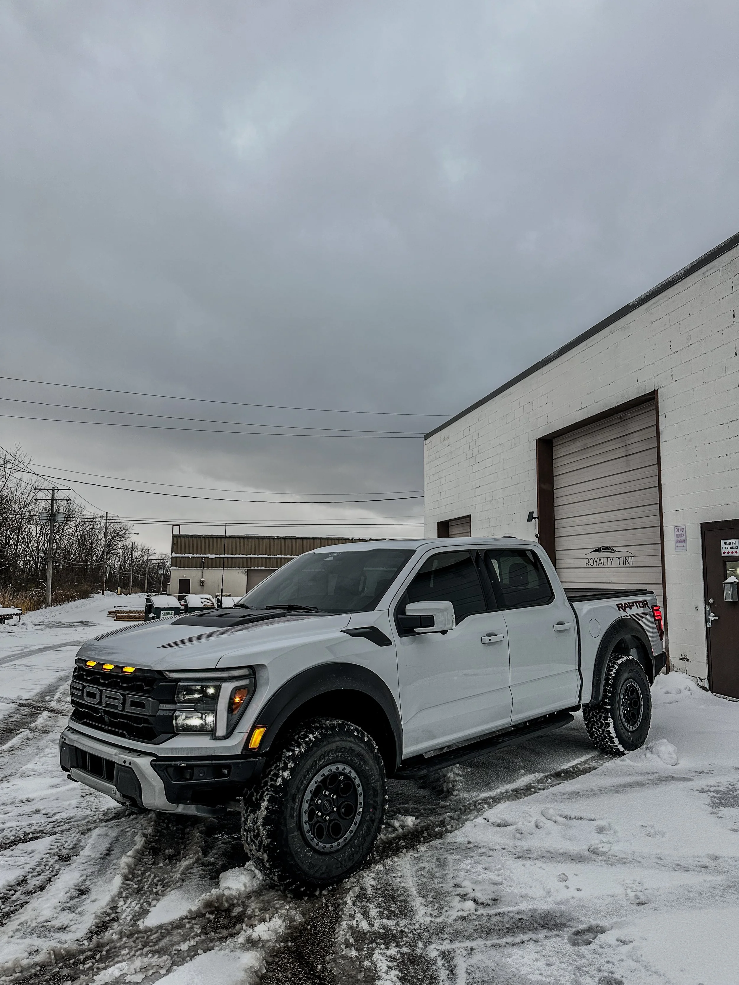 White Ford Raptor pickup truck parked on snow-covered ground outside a white brick building with a large garage door and a dark cloudy sky overhead.